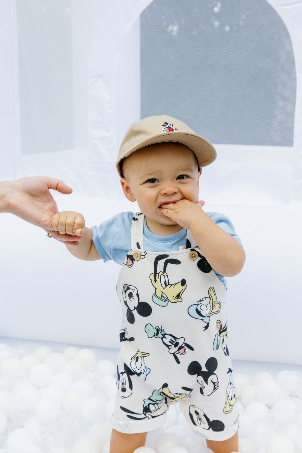 Cute baby boy in Mickey Mouse overalls and tan cap standing in white ball pit at Disney themed first birthday party.