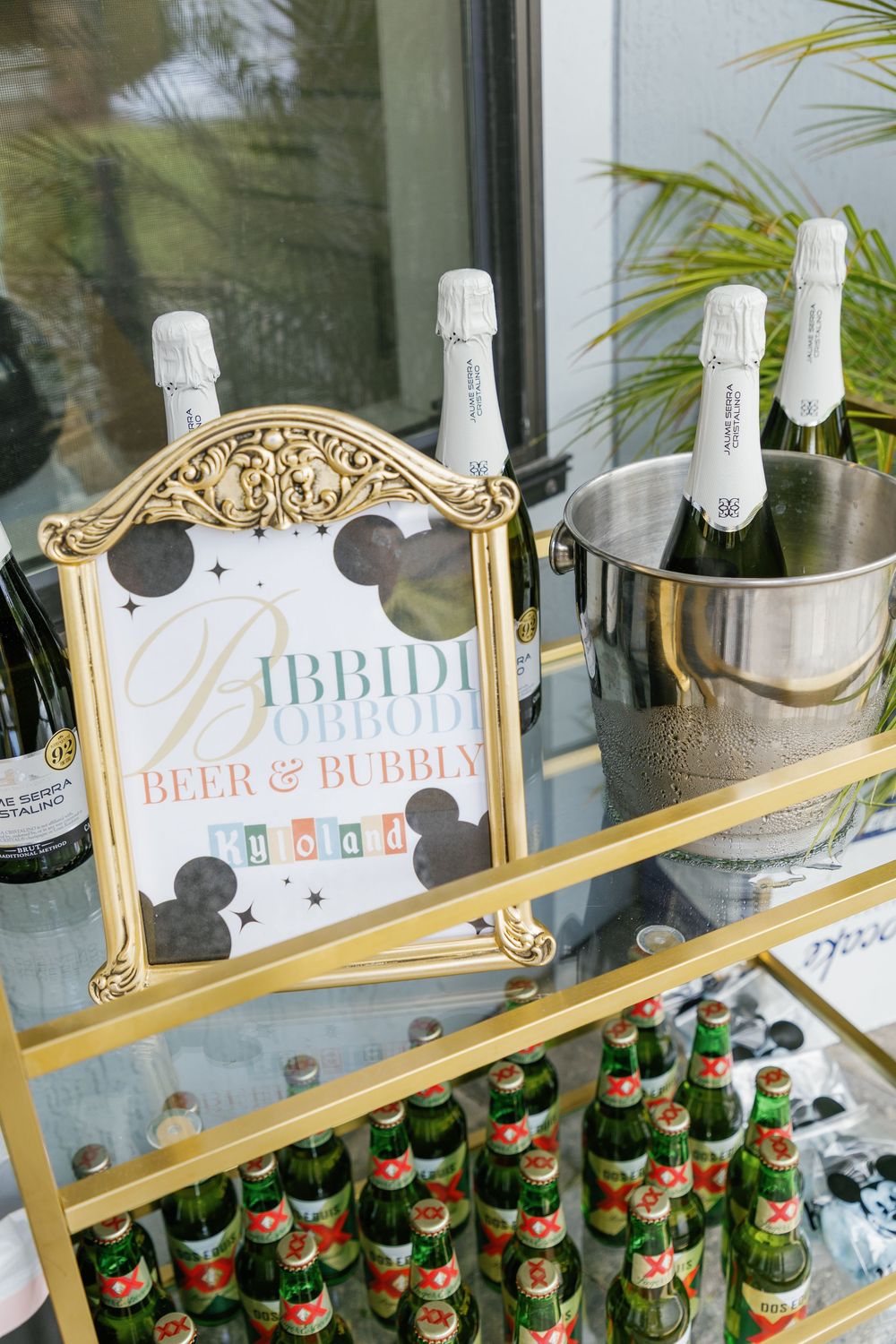 Gold bar cart with Bibbidi Bobbidi Beer and Bubbly sign, champagne bottles, and green beer bottles at Disney party.