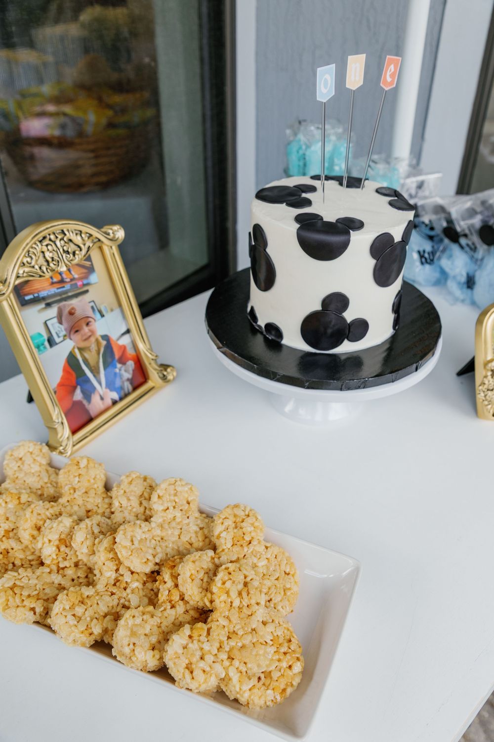 Mickey Mouse first birthday cake in black and white with Rice Krispie treats and framed baby photo on dessert table.