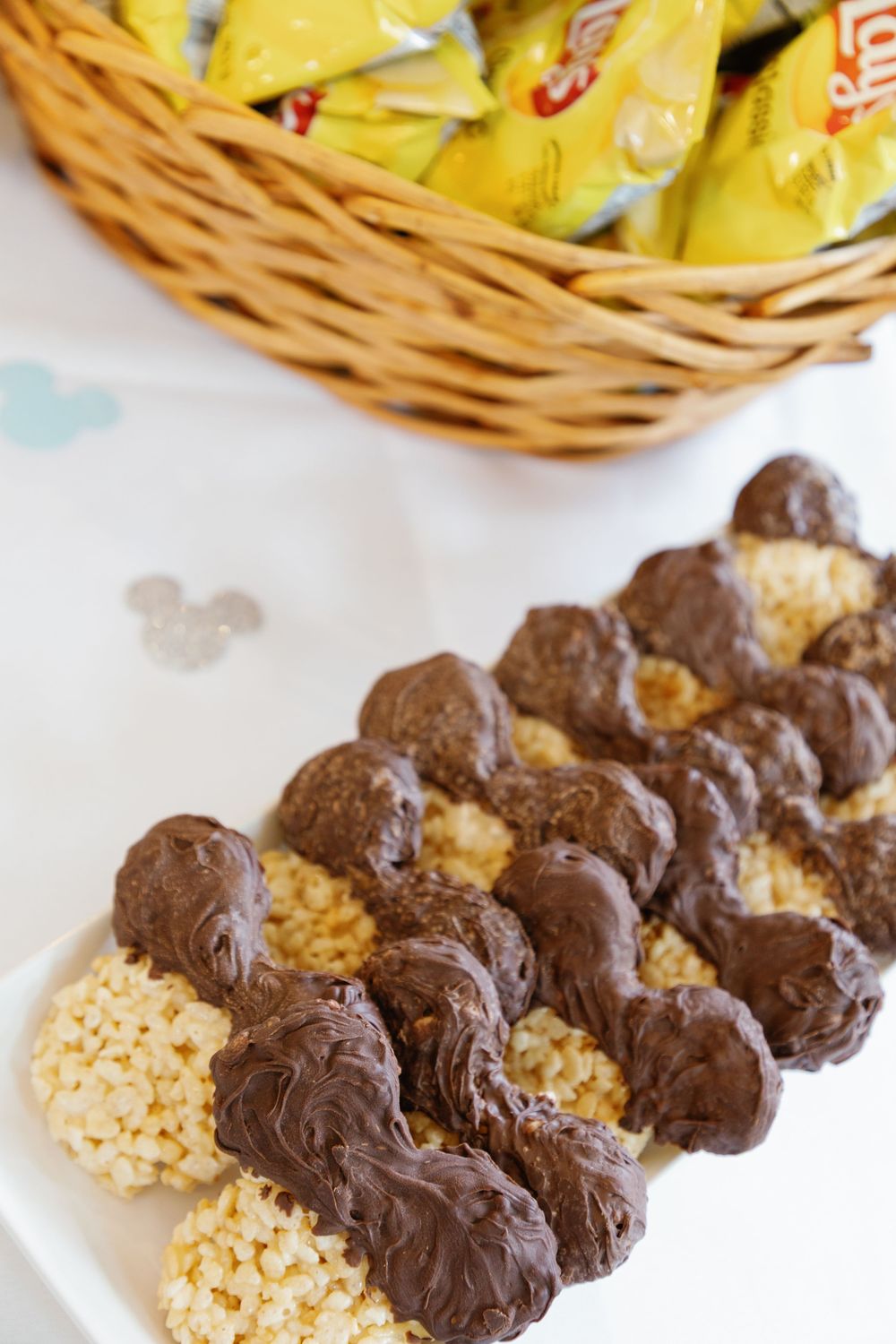 Mickey Mouse shaped Rice Krispie treats dipped in chocolate displayed on plate with candy basket at Disney party.