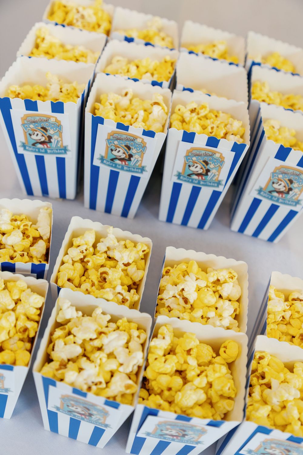 Blue and white striped Mickey Mouse themed popcorn boxes filled with buttered popcorn arranged on a table.