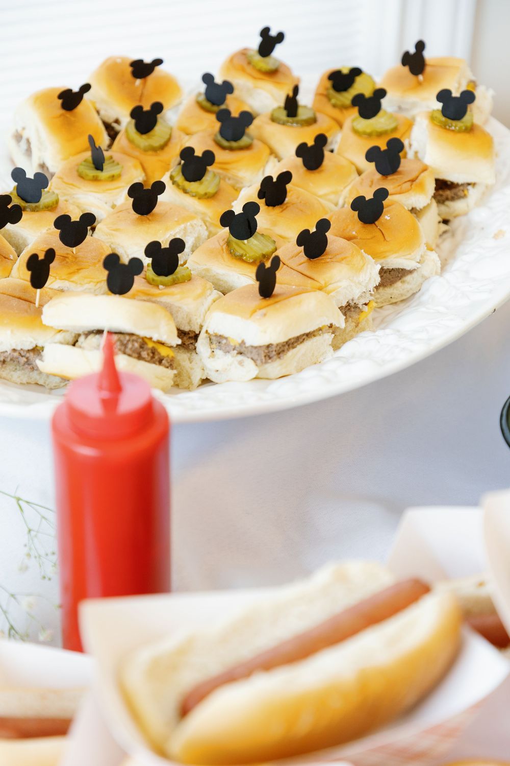 Mini Mickey Mouse slider burgers with black Mickey ear picks on tiered platter with ketchup and hot dogs.