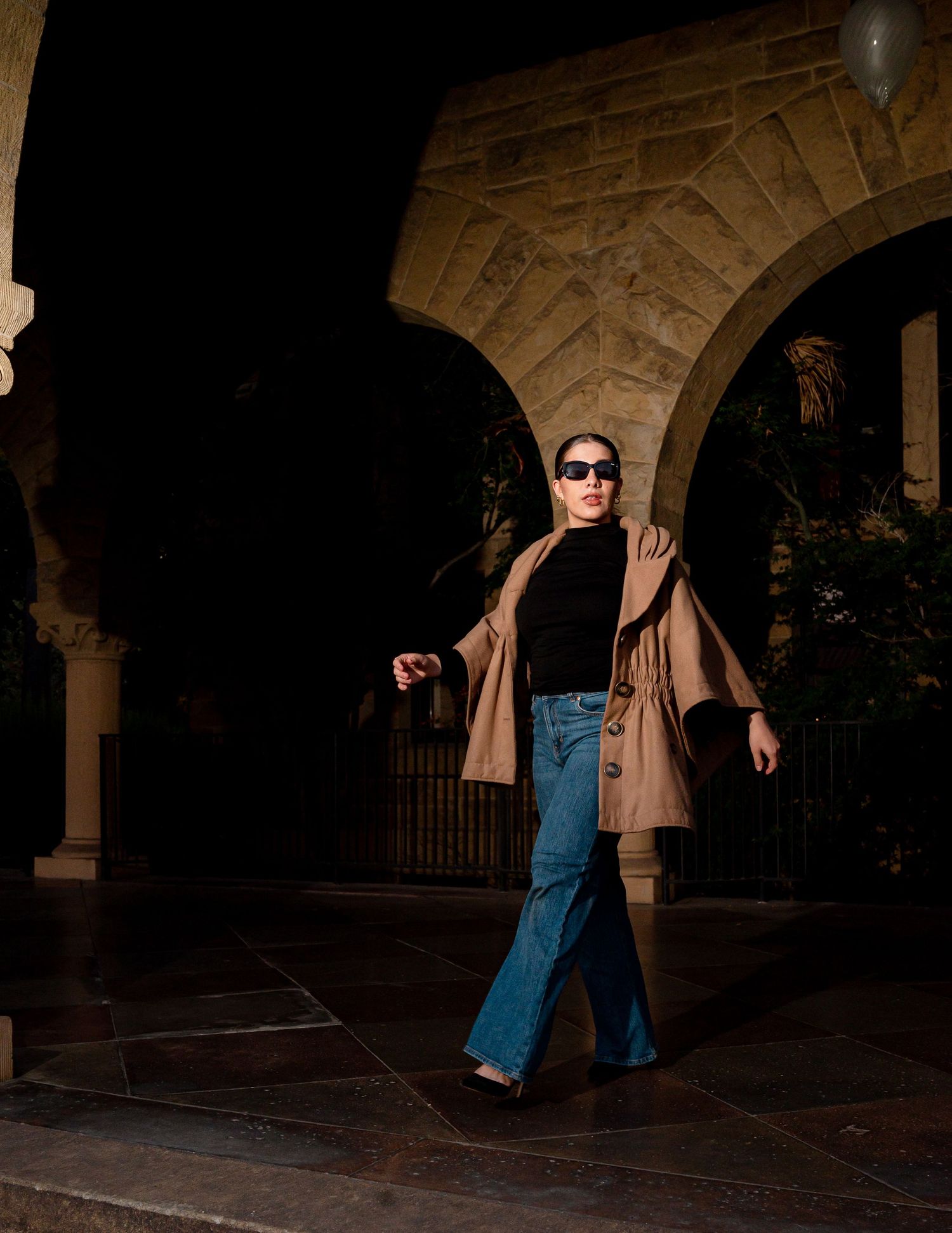 A person wearing sunglasses, brown coat and jeans walks confidently through an arched stone passageway at dusk.