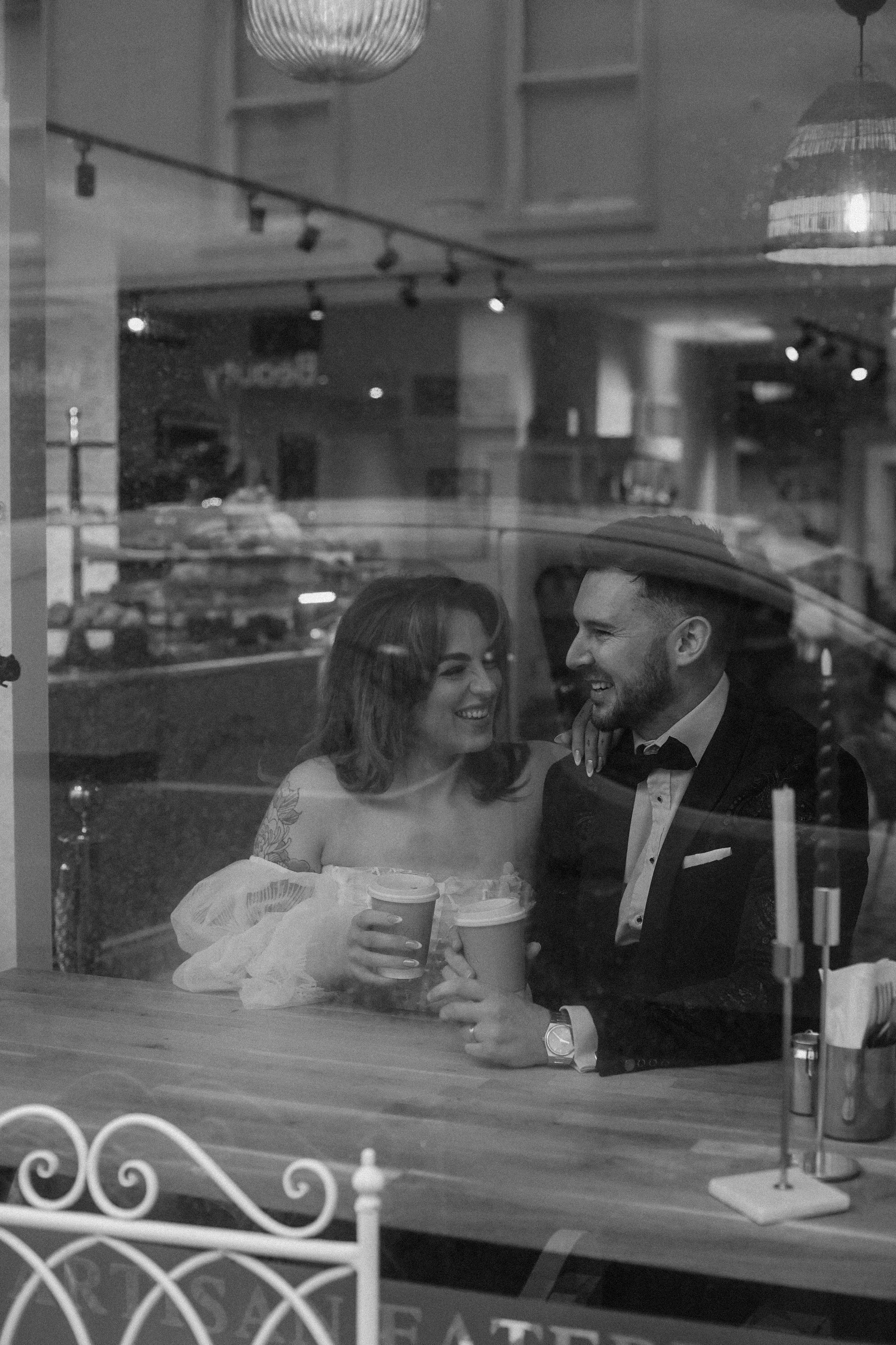 A black and white photo of a well-dressed couple sharing drinks at a romantic restaurant table by the window.