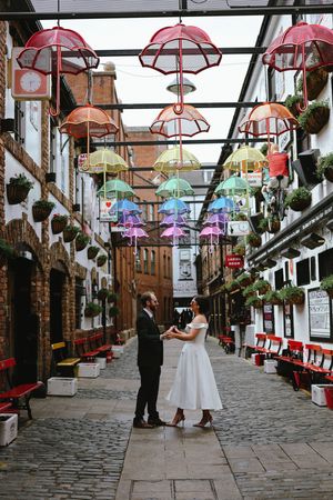 Two people stand in a charming alleyway decorated with colorful umbrella installations and brick walls in an urban setting.