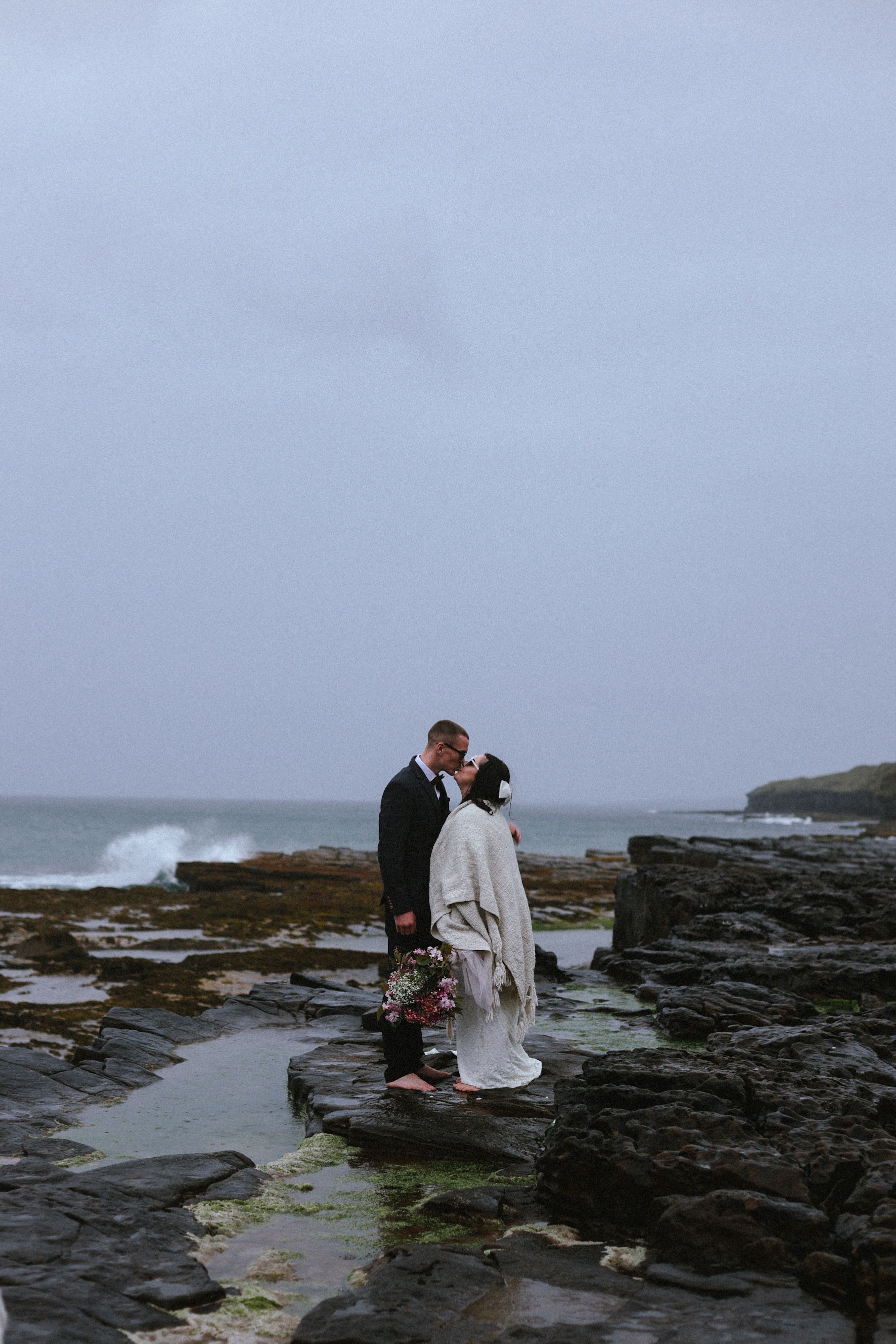 Two newlyweds share a romantic moment on rugged coastal rocks as waves crash against the shore on an overcast day.