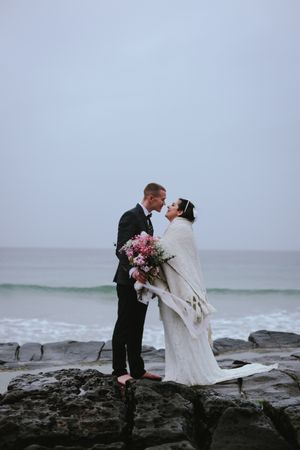 A romantic wedding photoshoot on dramatic rocky coastline with crashing waves in the background.