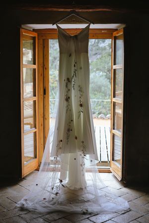 A white wedding dress hangs elegantly in front of open wooden French doors with natural light streaming through.
