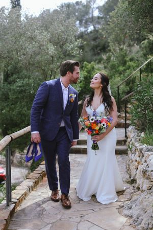 Happy newlyweds share a joyful moment on a rustic stone pathway while holding a vibrant wedding bouquet.