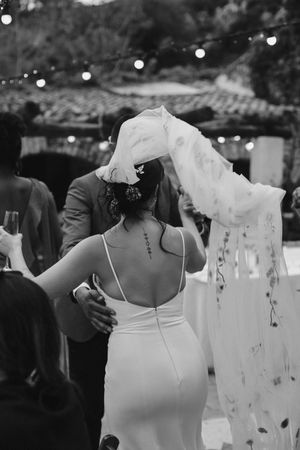 Black and white photo of a couple embracing during a wedding reception with twinkle lights and veil in the background.