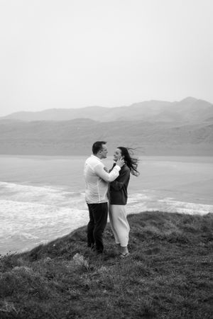 A romantic couple embraces on a scenic cliff overlooking a misty coastal landscape in black and white.