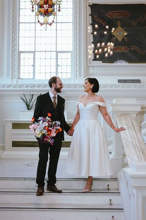 A wedding couple stands on elegant white marble stairs in a grand historic venue with stained glass windows.