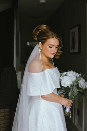 A bride in an off-shoulder white lace wedding dress holds a bouquet of white flowers while wearing a long flowing veil.