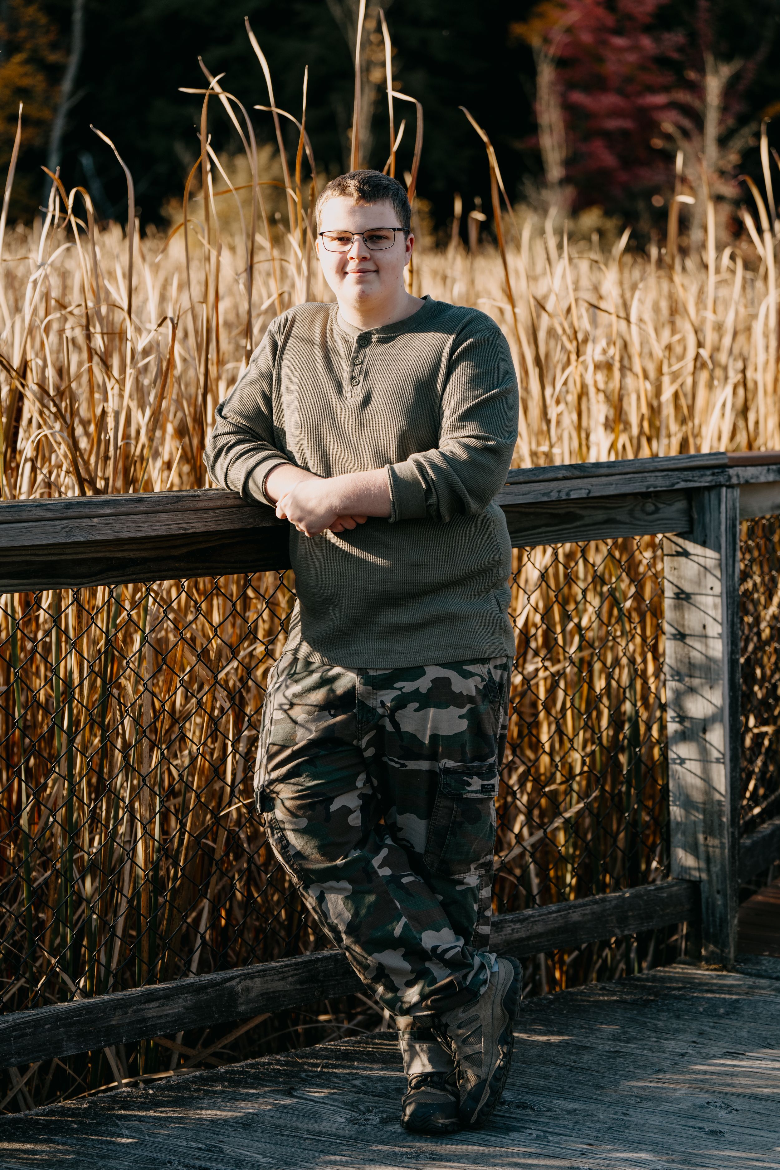 Someone in a beige sweater and camouflage pants leans casually against a wooden railing with tall reeds in the background.