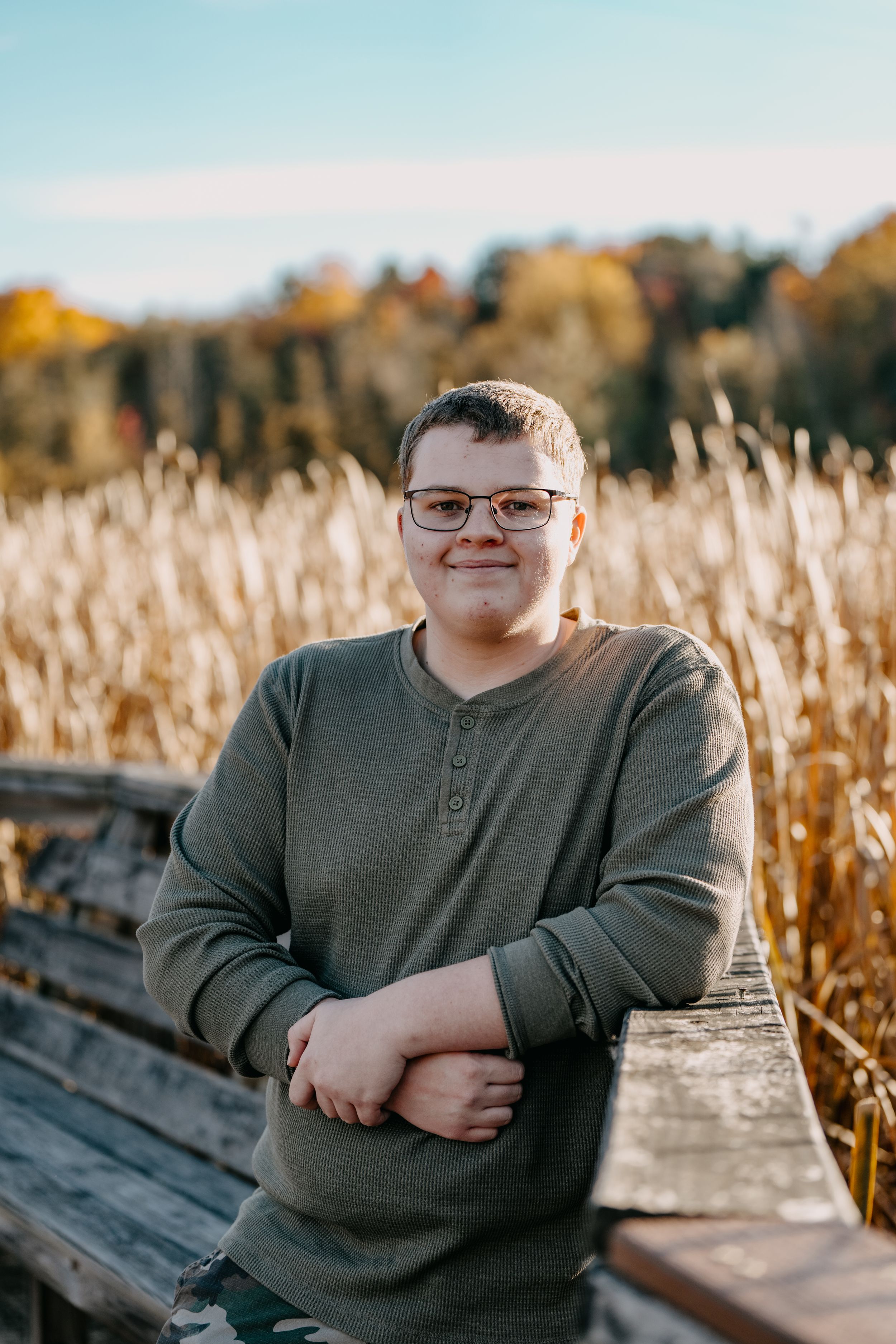 Someone wearing glasses and an olive henley sweater poses at sunset on a wooden boardwalk surrounded by fall foliage.