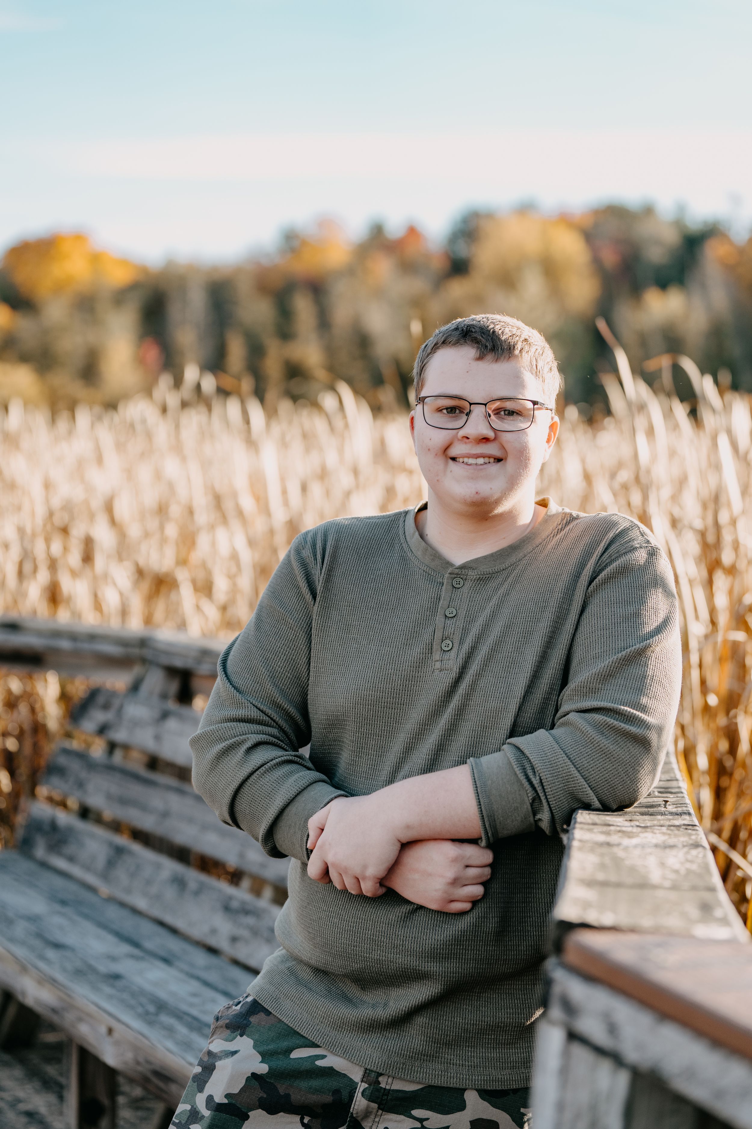 A person in a gray henley sweater stands smiling on a wooden dock surrounded by autumn cattails at sunset.