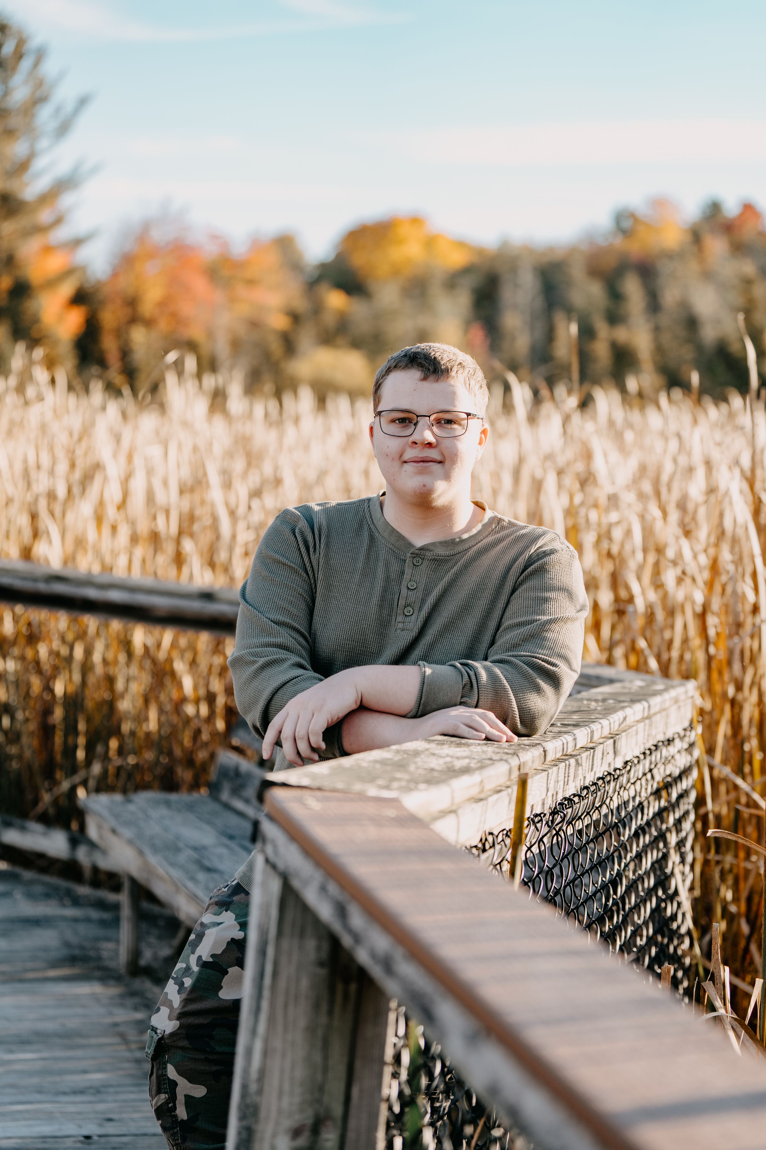 Portrait photograph of someone wearing glasses and a green henley sweater leaning on a wooden boardwalk railing amid autumn reeds.