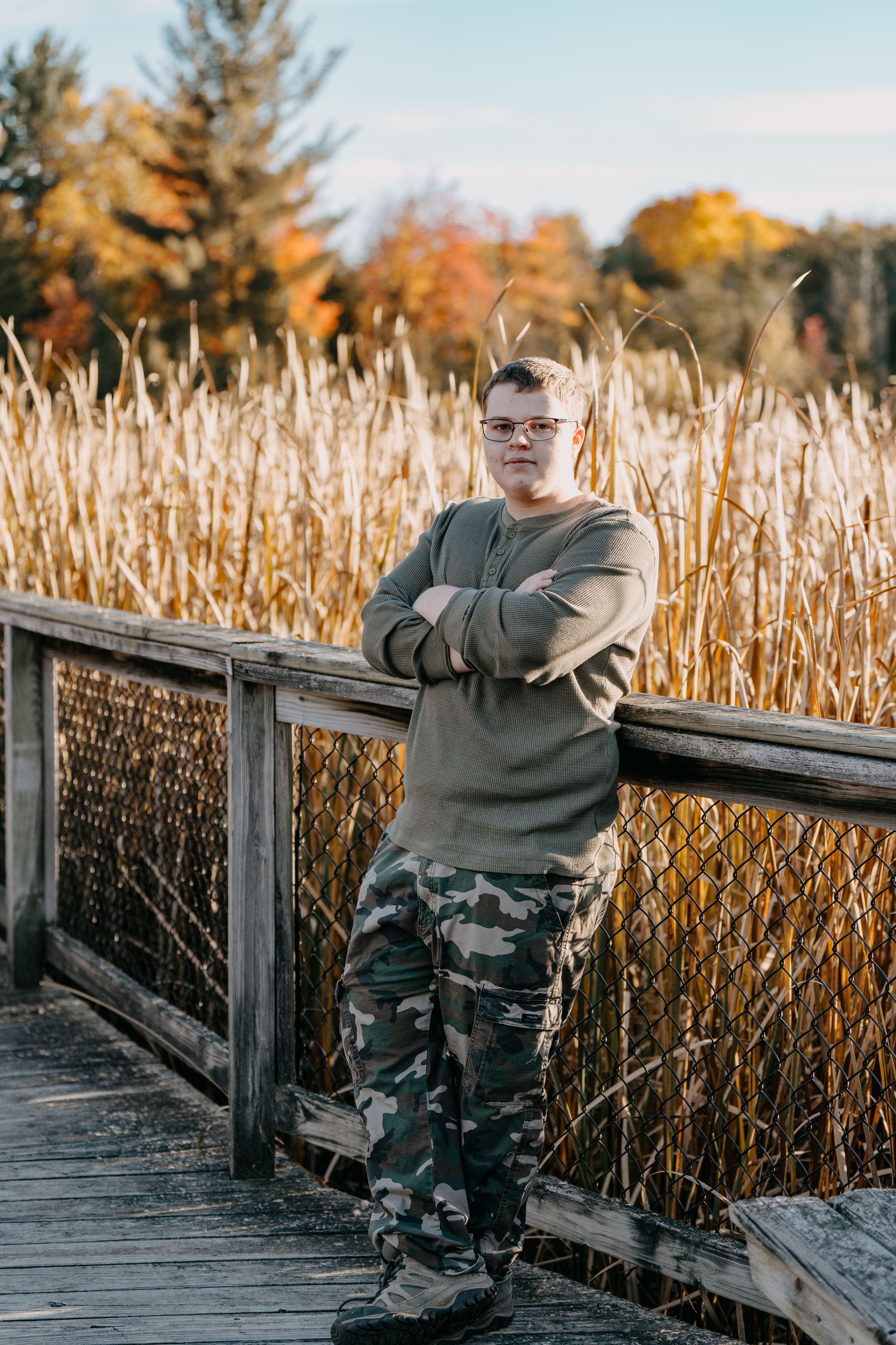 A person in camo pants and olive sweater stands on a wooden boardwalk surrounded by tall autumn cattails at sunset.