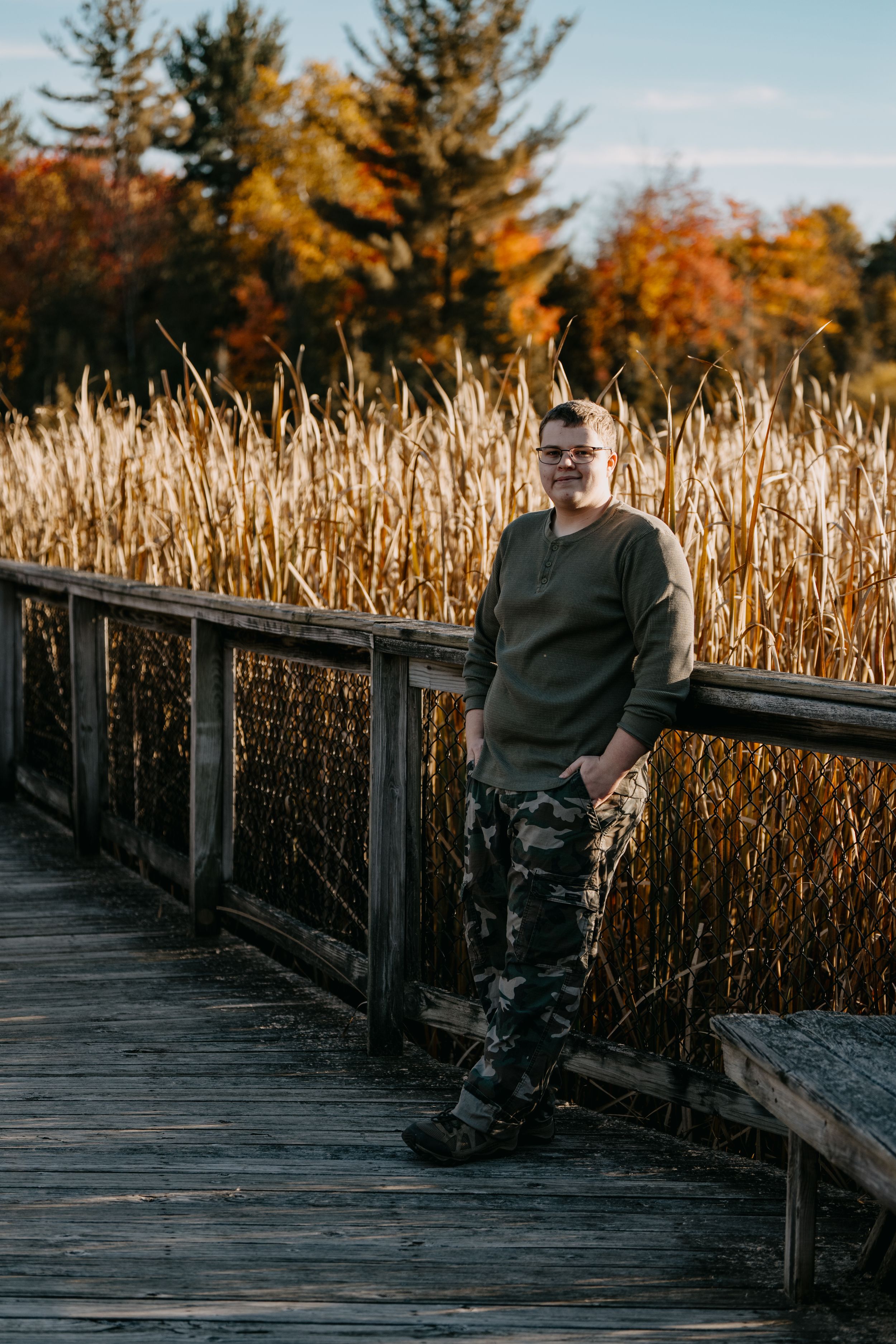 Someone stands on a wooden boardwalk surrounded by tall golden cattails and autumn trees at sunset.
