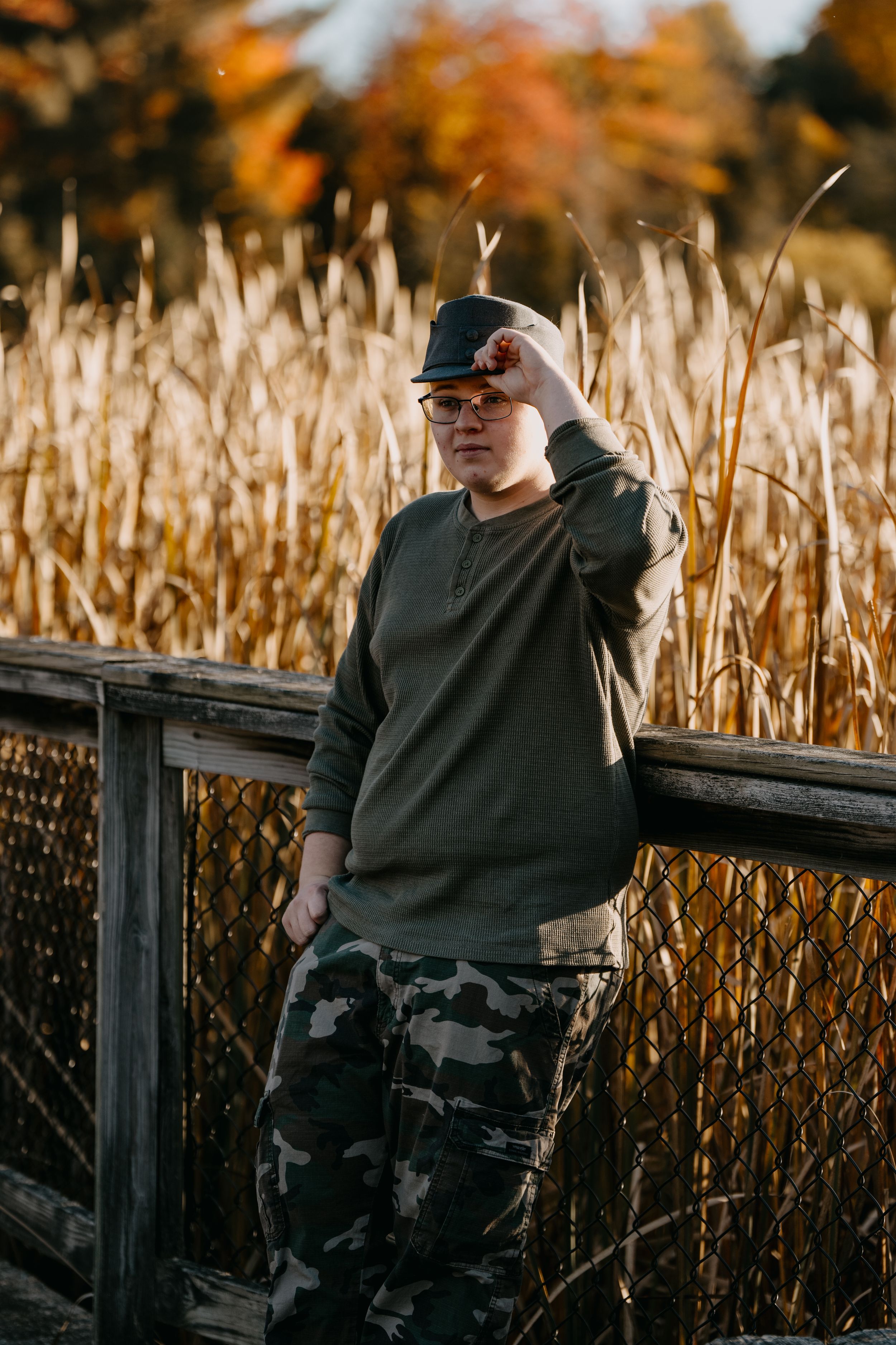 Figure in camouflage pants and dark sweater stands by wooden railing amongst tall autumn reeds at golden hour.