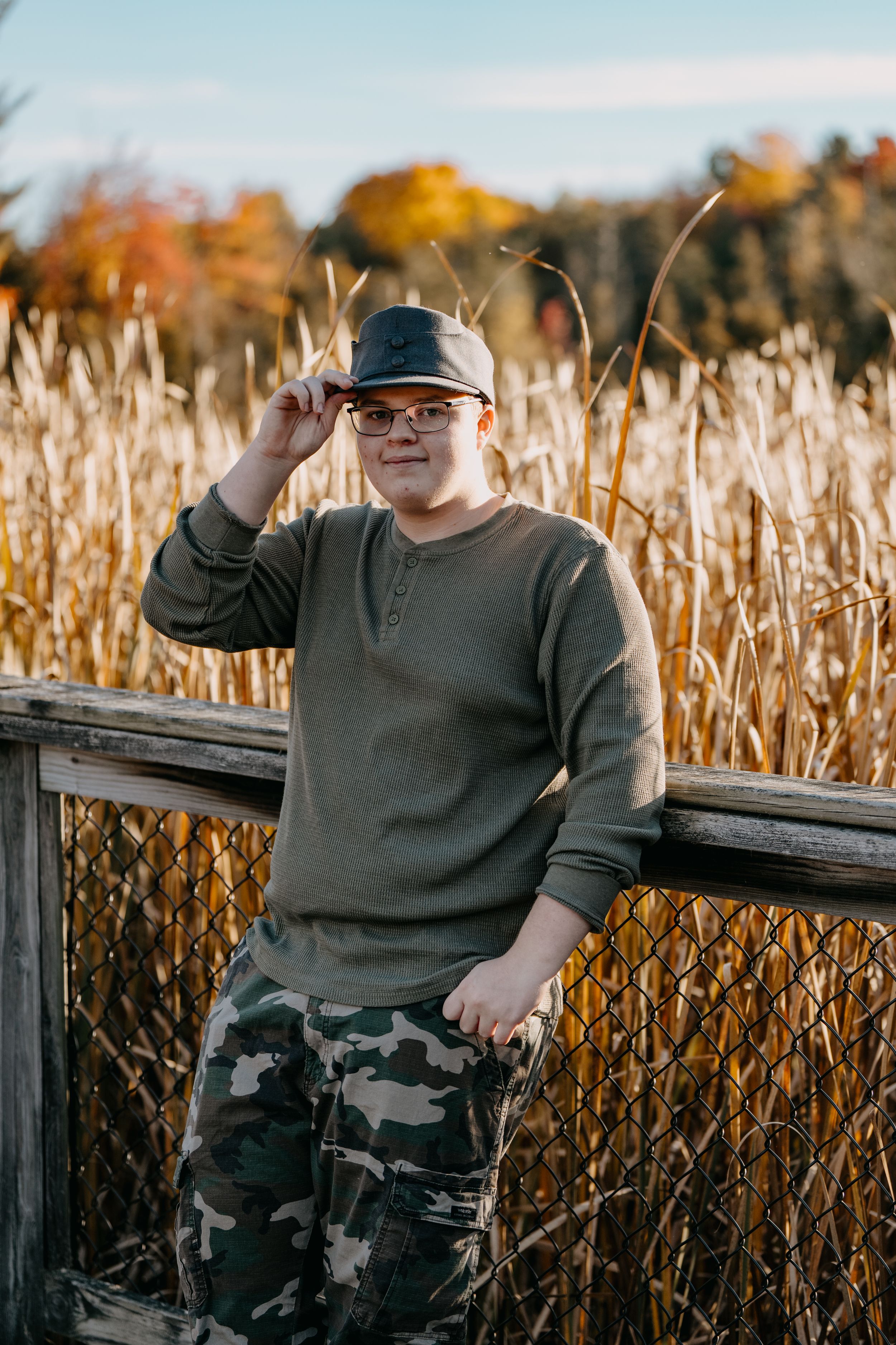 Someone in a casual olive sweater and camo pants poses near tall autumn grass and wooden railing at sunset.