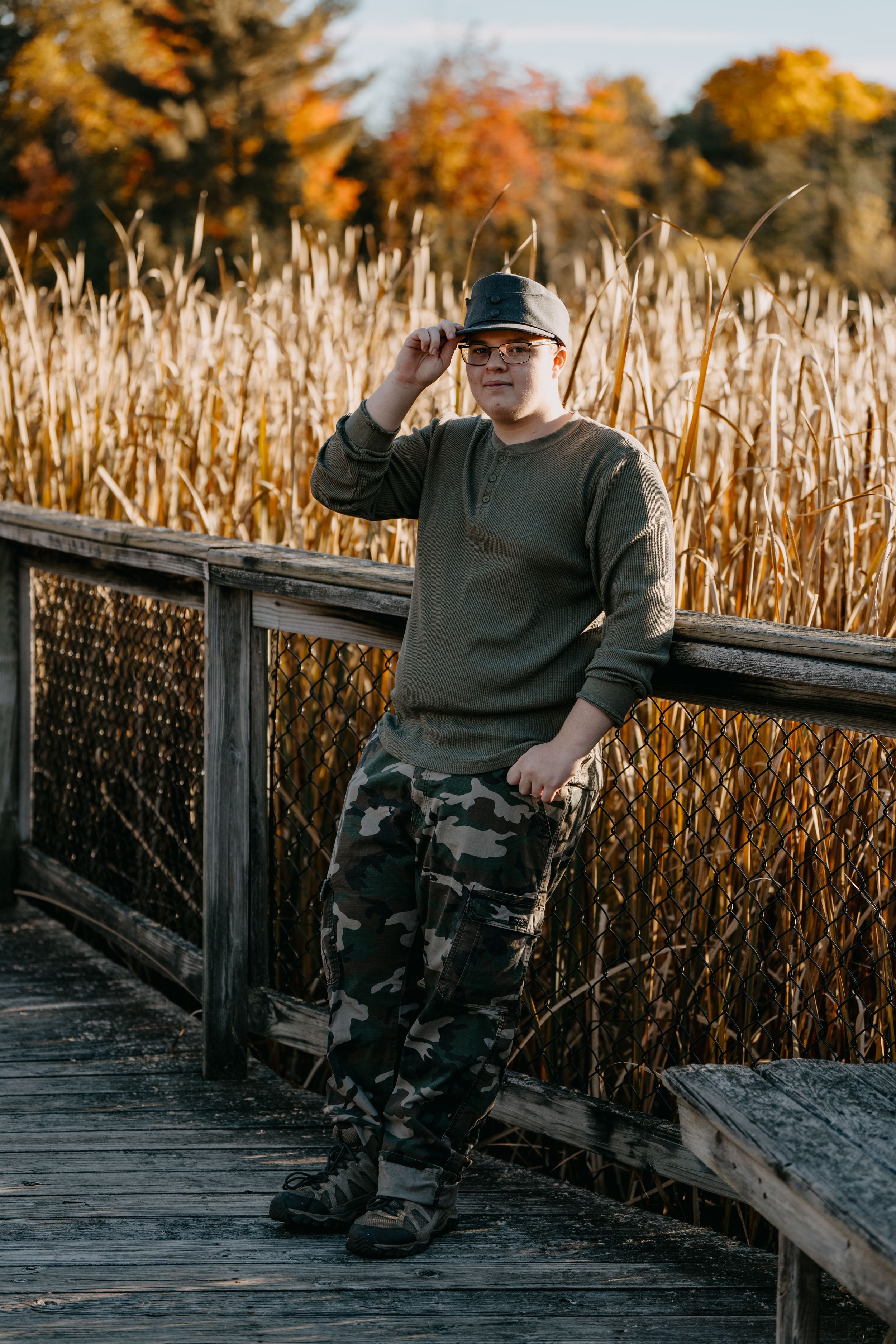 Person in olive sweater and camo pants poses against autumn backdrop of golden reeds and trees on wooden boardwalk.