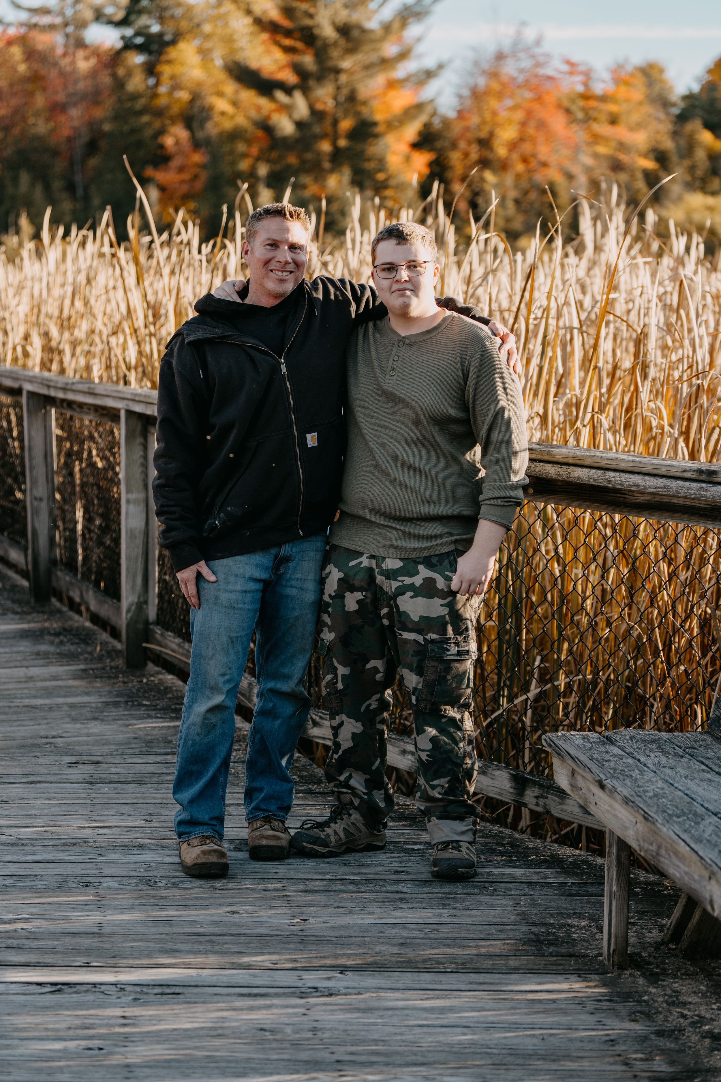 Two people stand together on a wooden boardwalk surrounded by autumn cattails and colorful fall foliage.