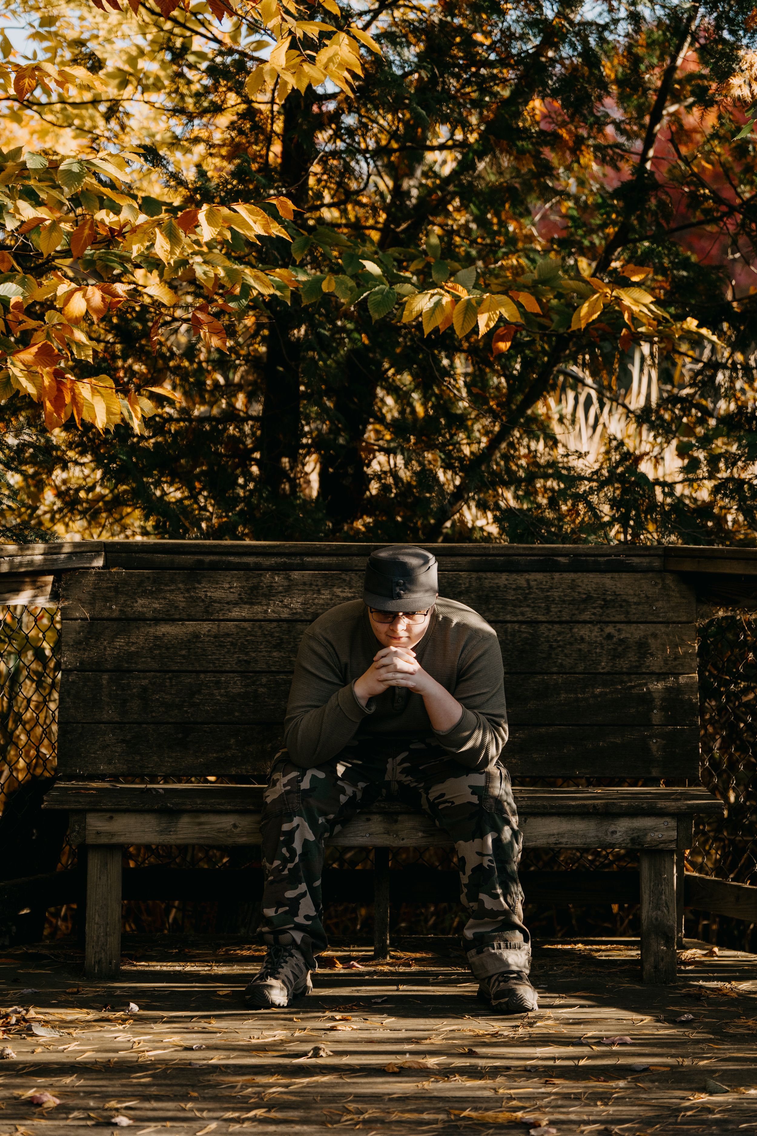 A solitary figure sits on a wooden bench surrounded by autumn foliage with golden leaves in a park setting.