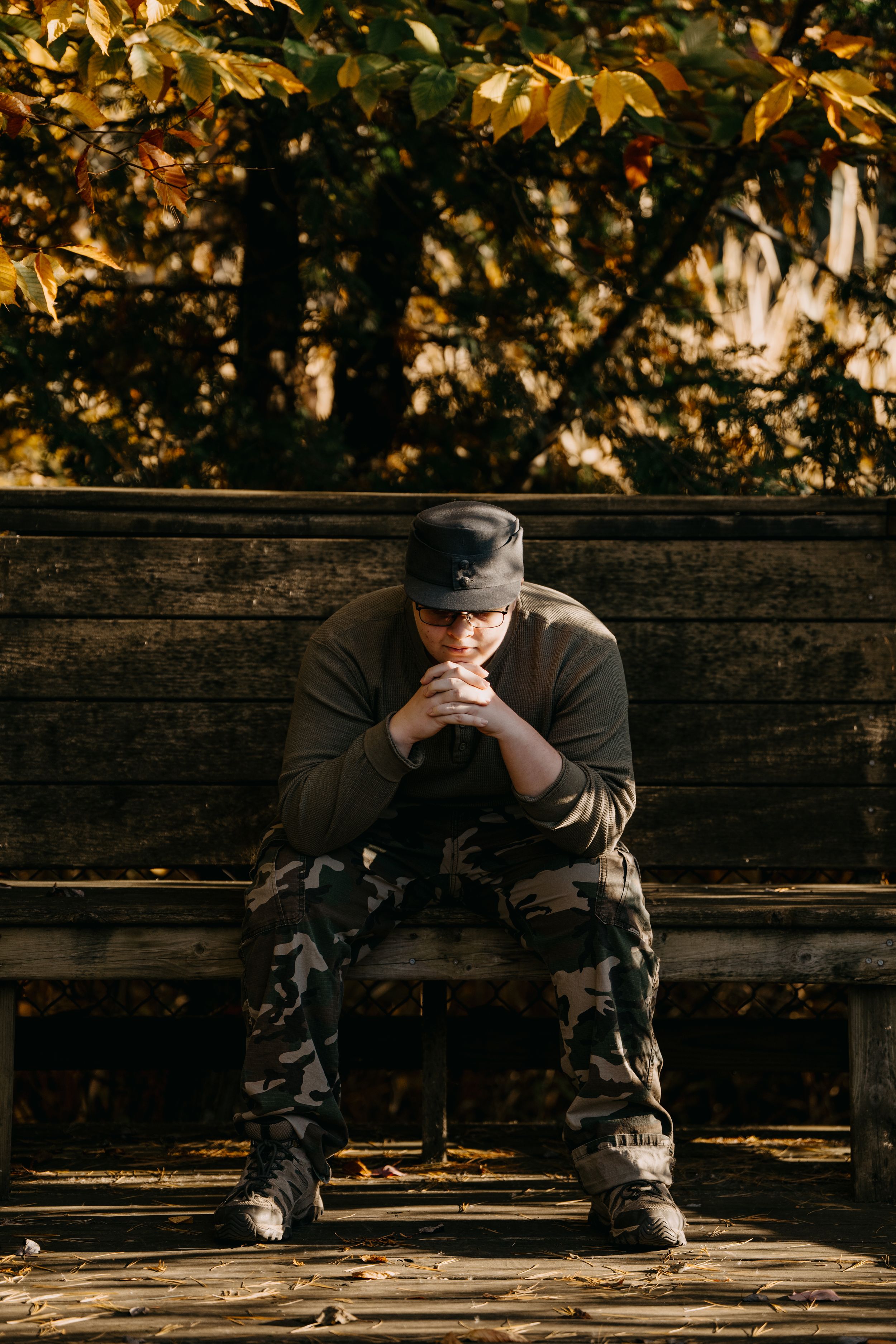 Person in camo pants and hat sits thoughtfully on rustic wooden bench surrounded by autumn foliage.
