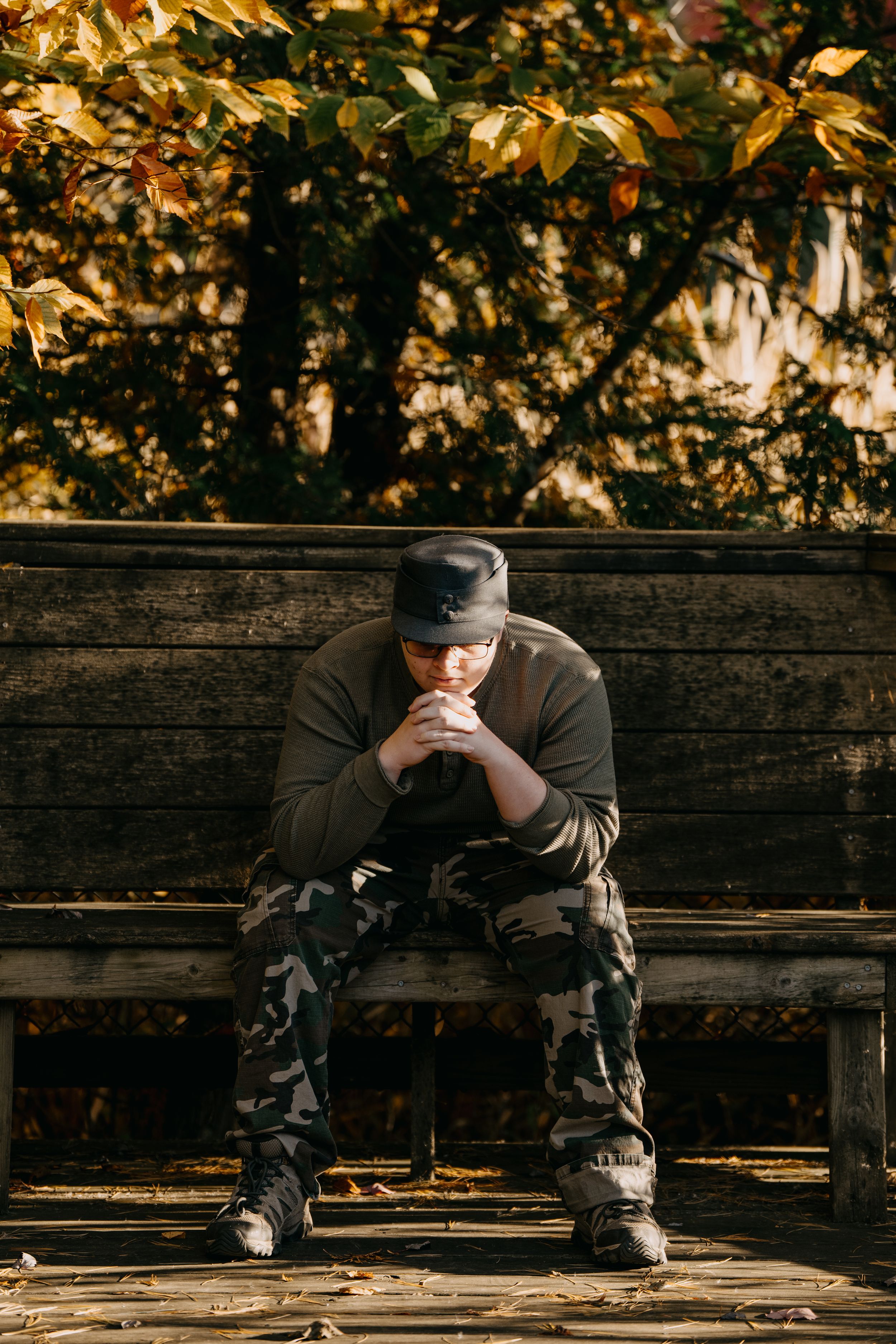 A person in camouflage pants and dark clothing sits pensively on a wooden bench during autumn.