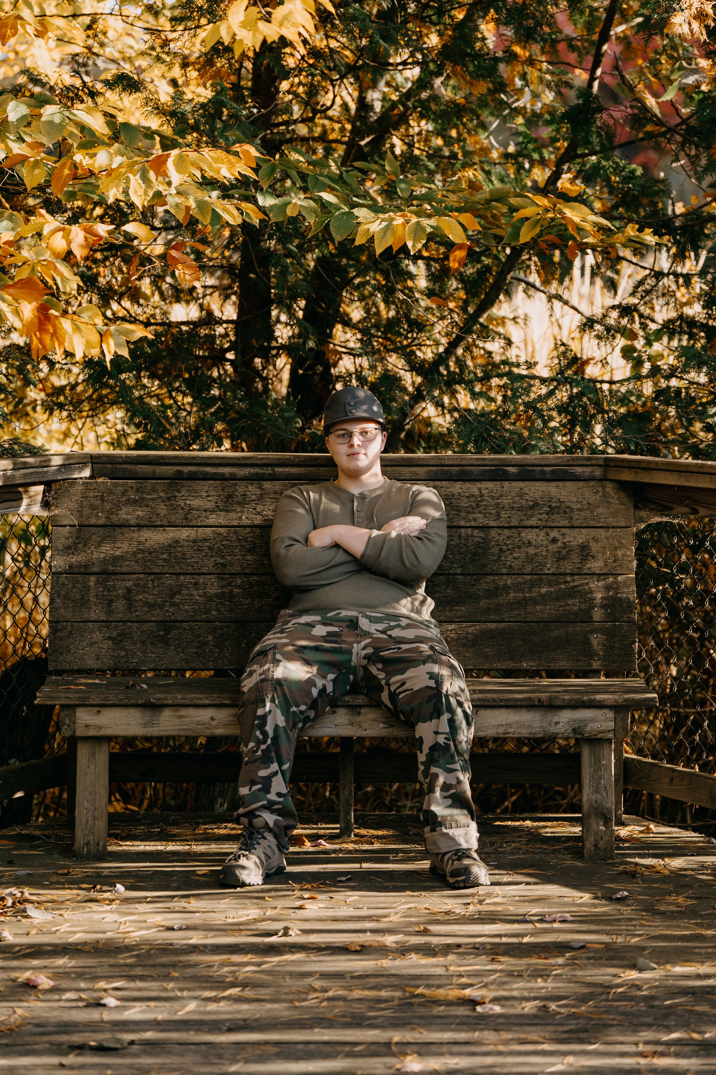 Someone wearing camouflage pants and a hat sits confidently on a wooden bench surrounded by autumn foliage.