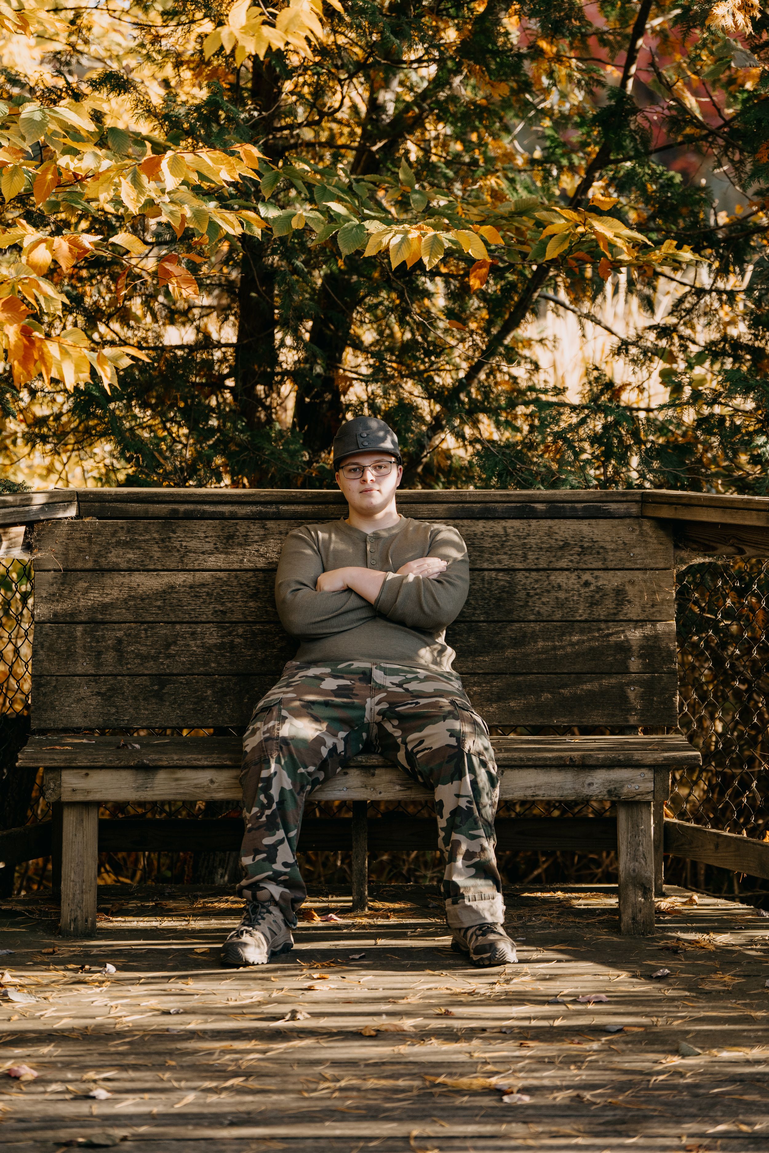 Person in casual military attire sits on rustic wooden bench surrounded by autumn foliage in outdoor setting.