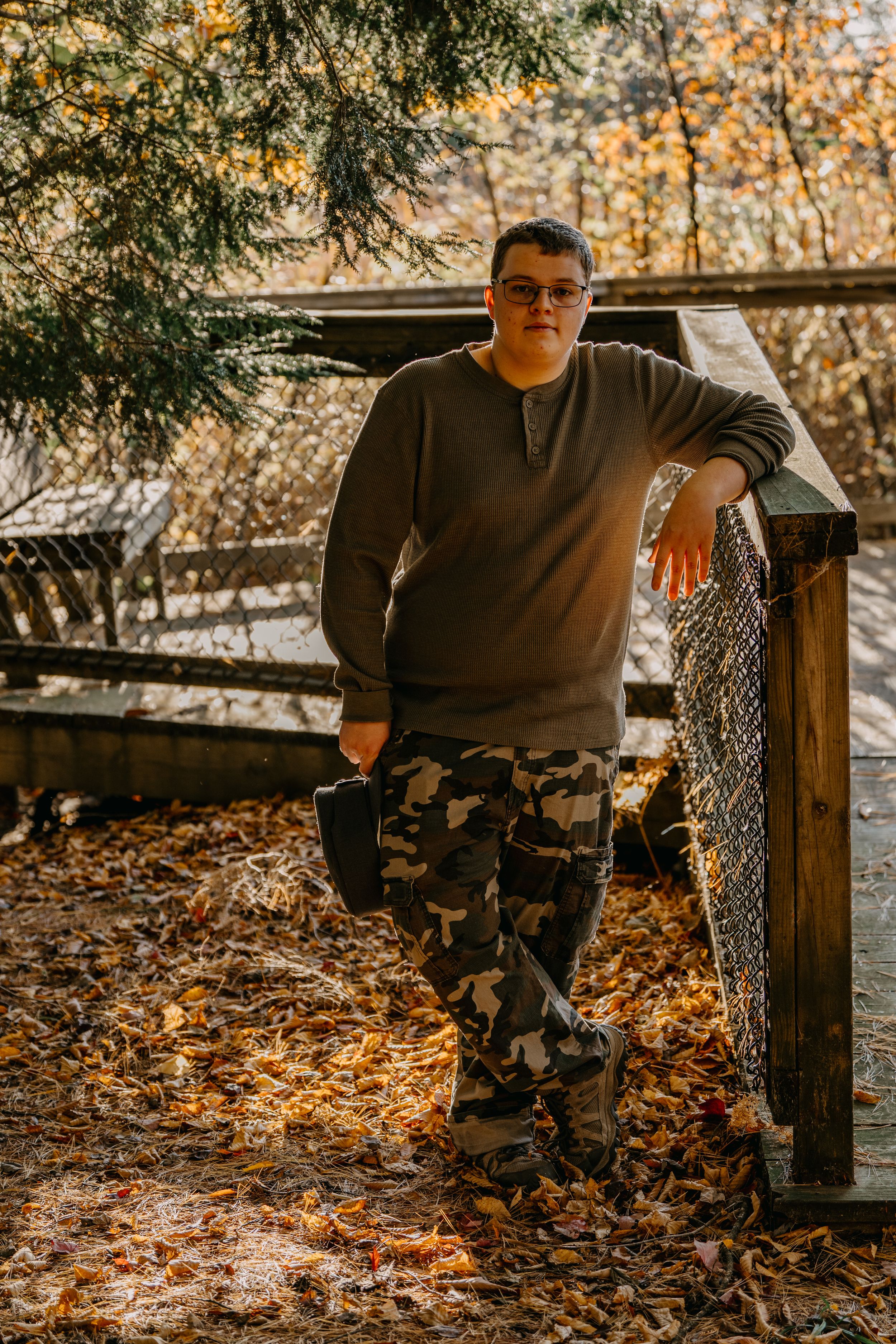 Person wearing grey sweater and camo pants leans against wooden fence in autumn forest setting.