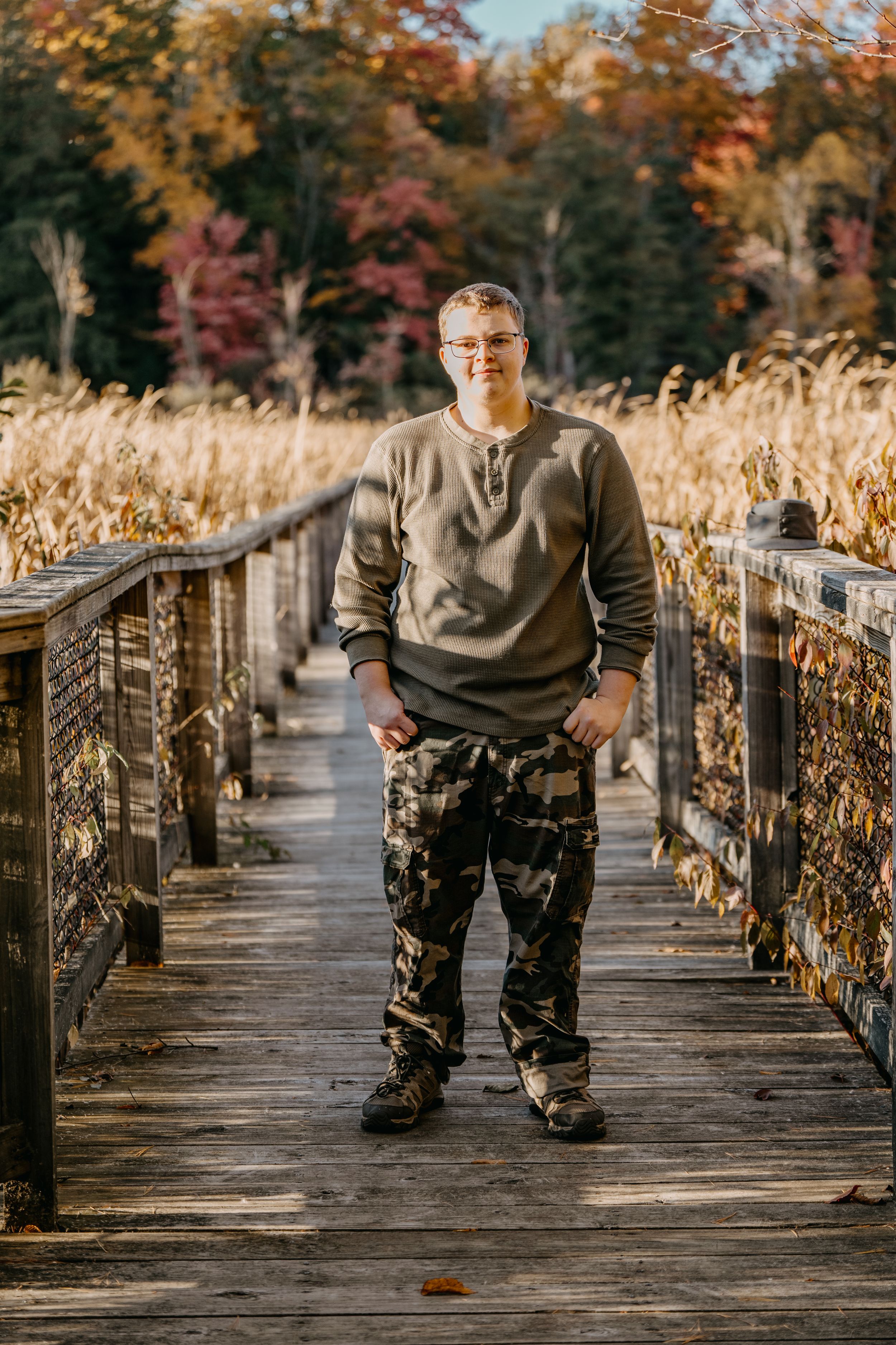Person in casual wear stands on rustic wooden walkway surrounded by fall foliage and dried corn stalks.