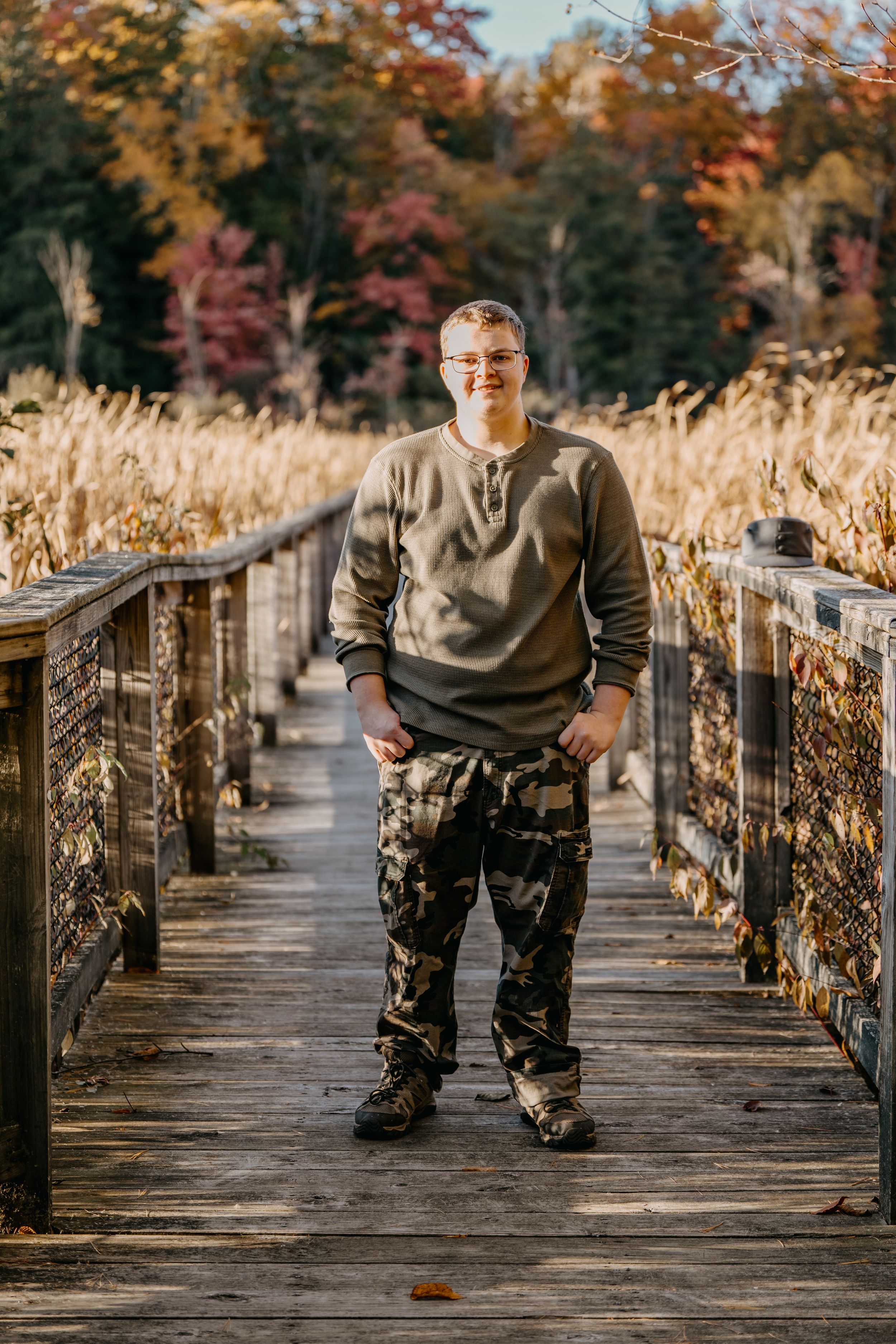 Person poses on a wooden boardwalk surrounded by tall autumn grasses and colorful fall trees in nature setting.