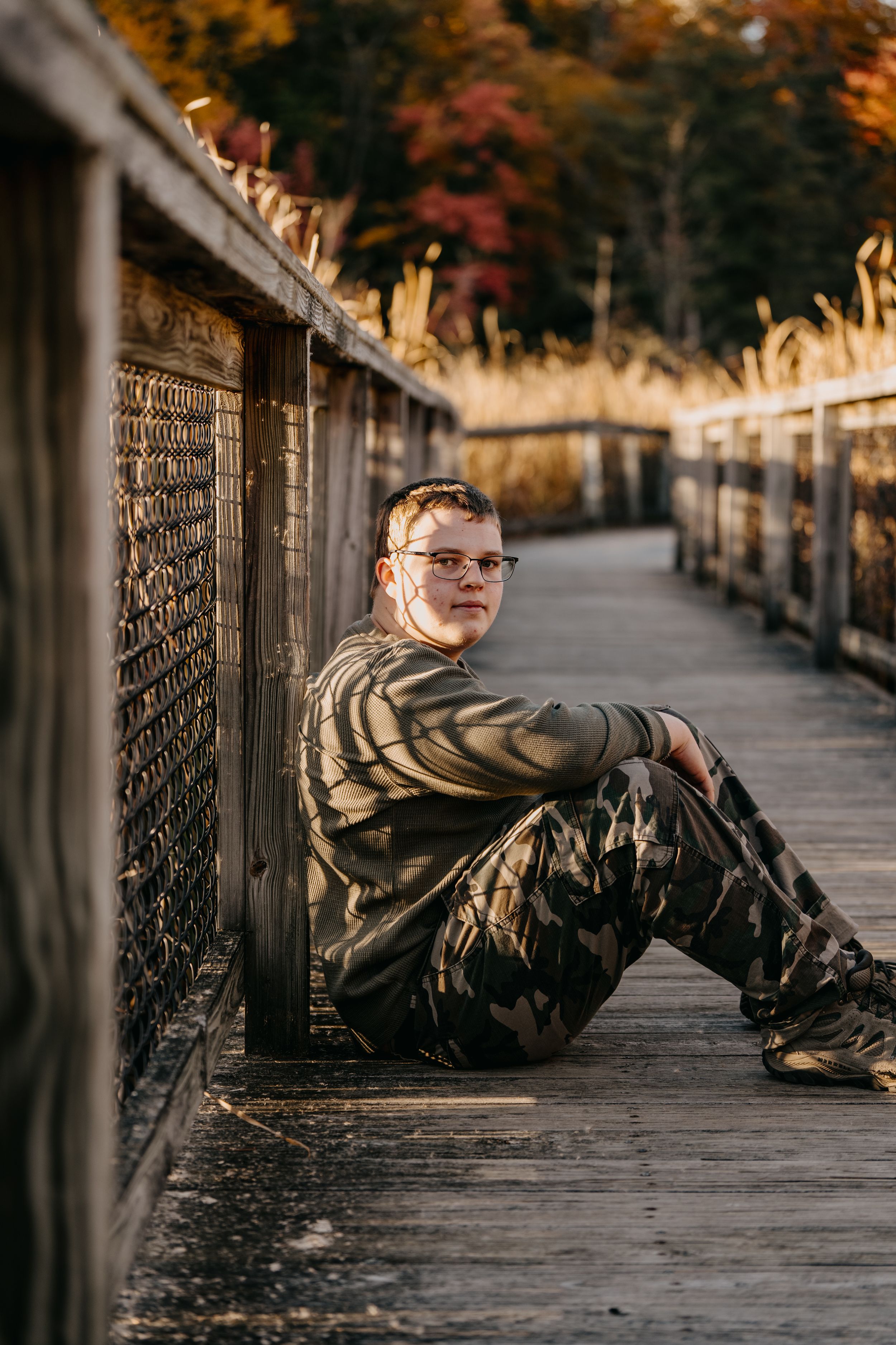 A person in camouflage clothing sits on a wooden bridge walkway with fall colors in the background.
