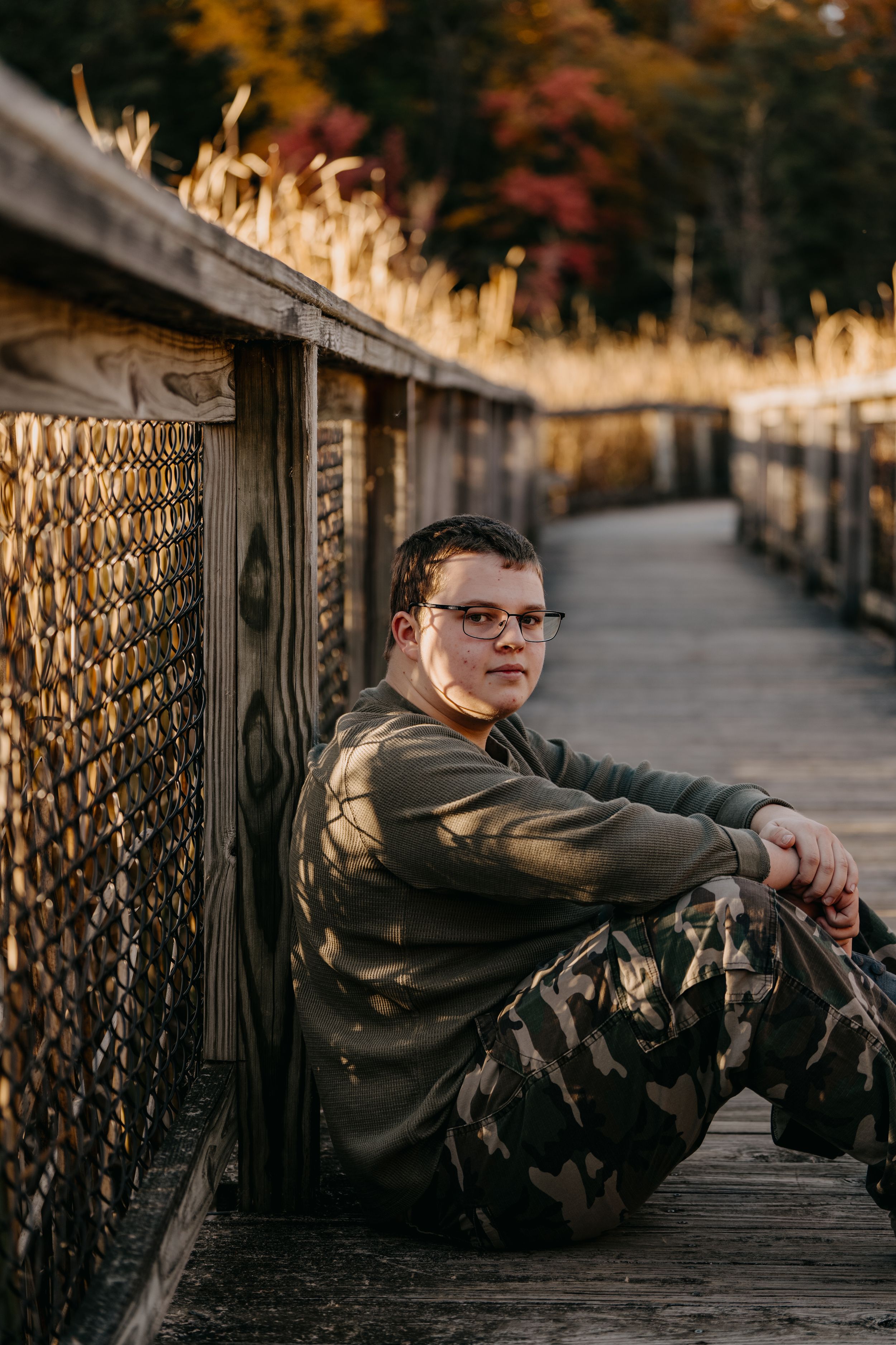 Person in casual attire sitting on wooden bridge walkway with fall colors in background.