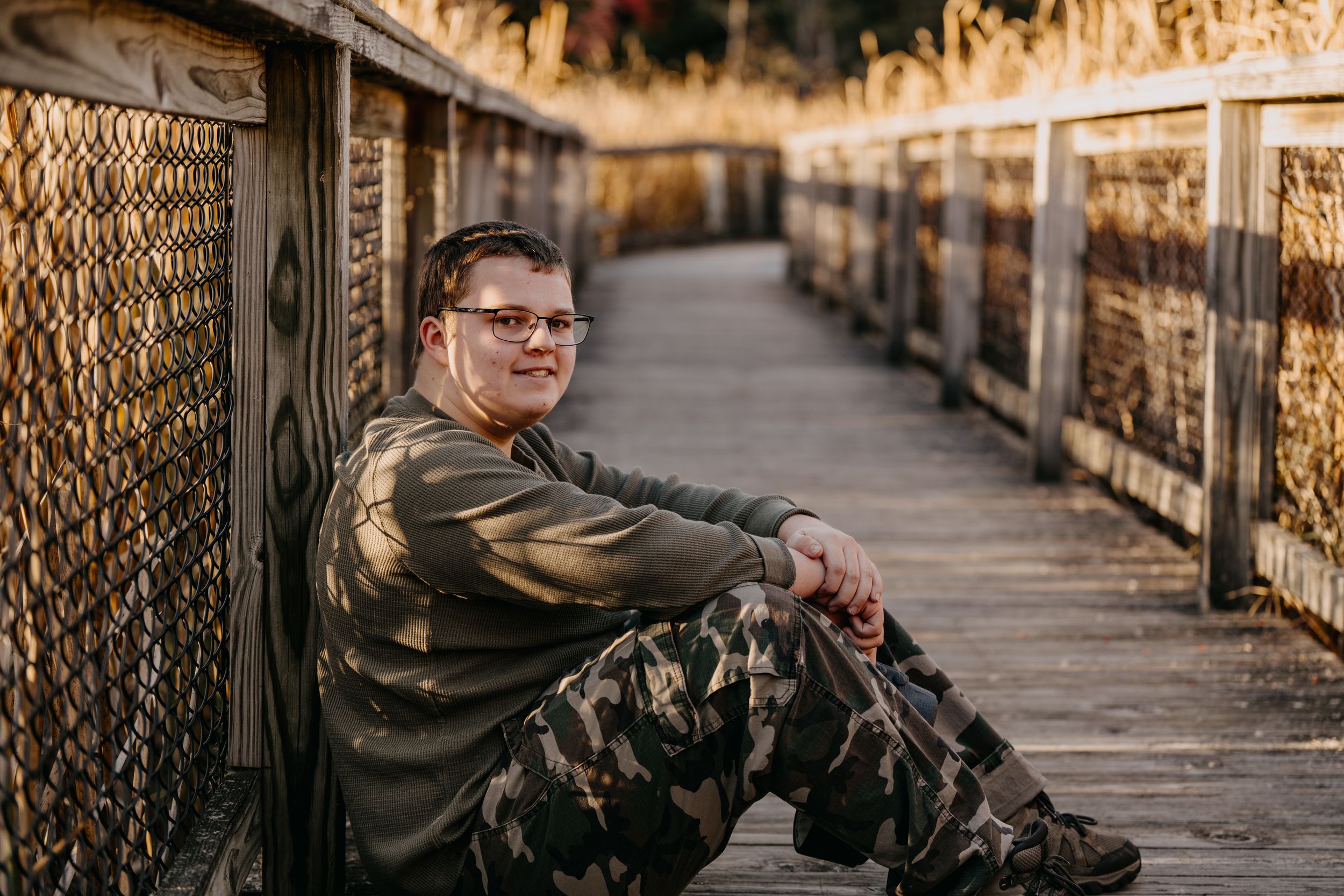 Someone wearing camouflage pants and a brown jacket sits on a wooden boardwalk with chain link fencing in autumn.