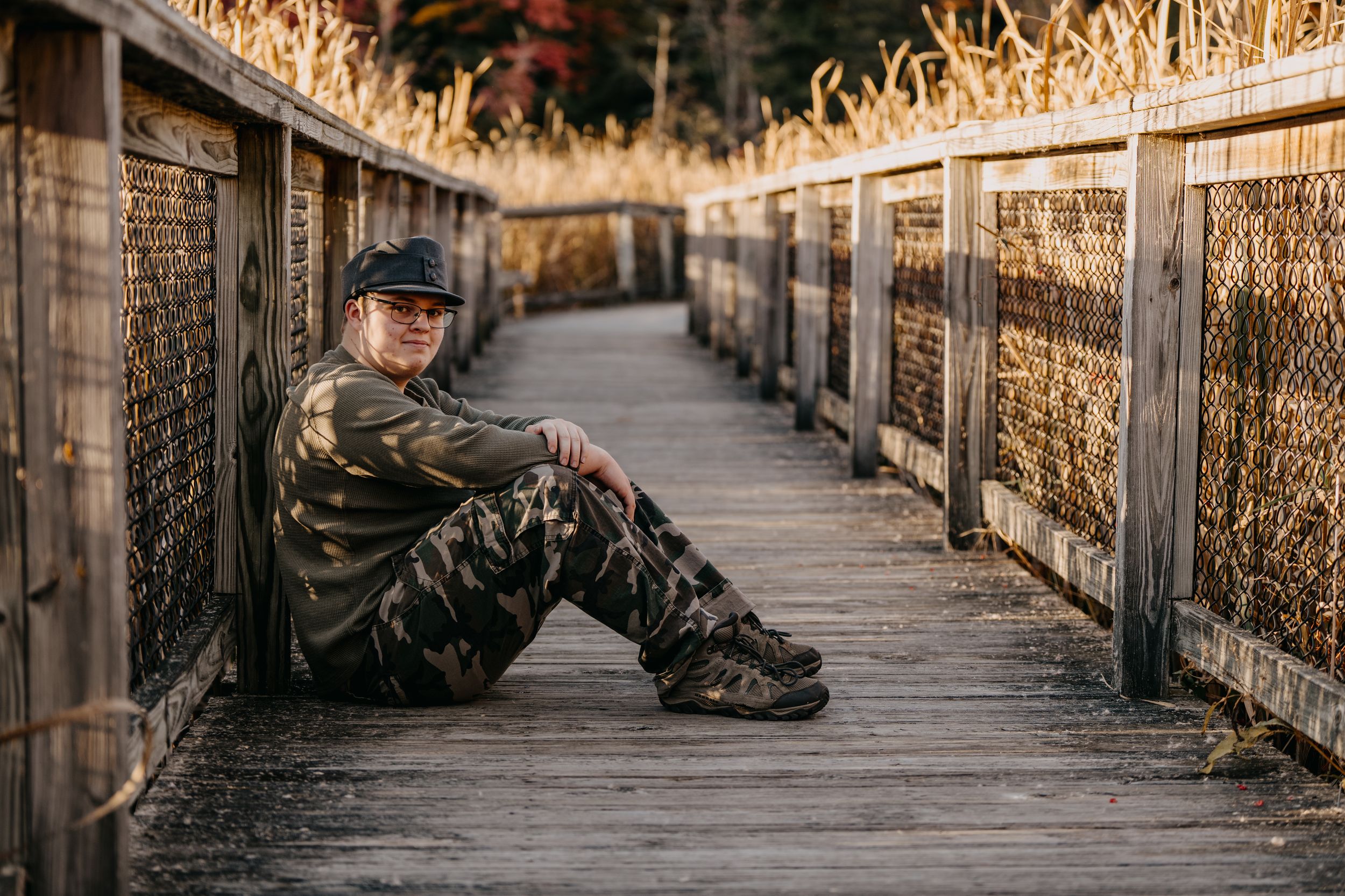 Person in camouflage clothing sitting on wooden boardwalk surrounded by tall dry grass at sunset.