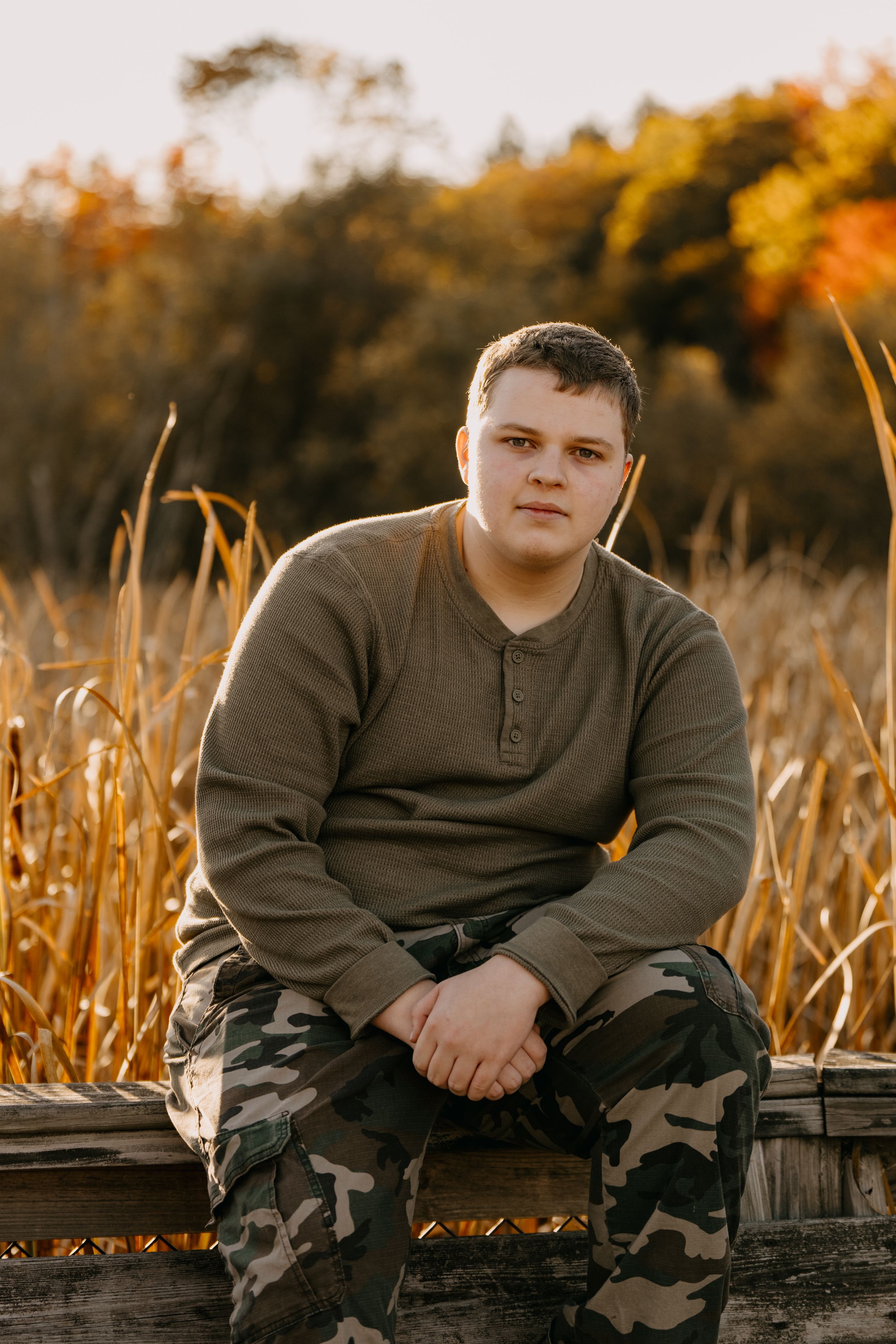 Portrait of a young person in brown henley and camo pants sitting on wooden dock during autumn sunset.