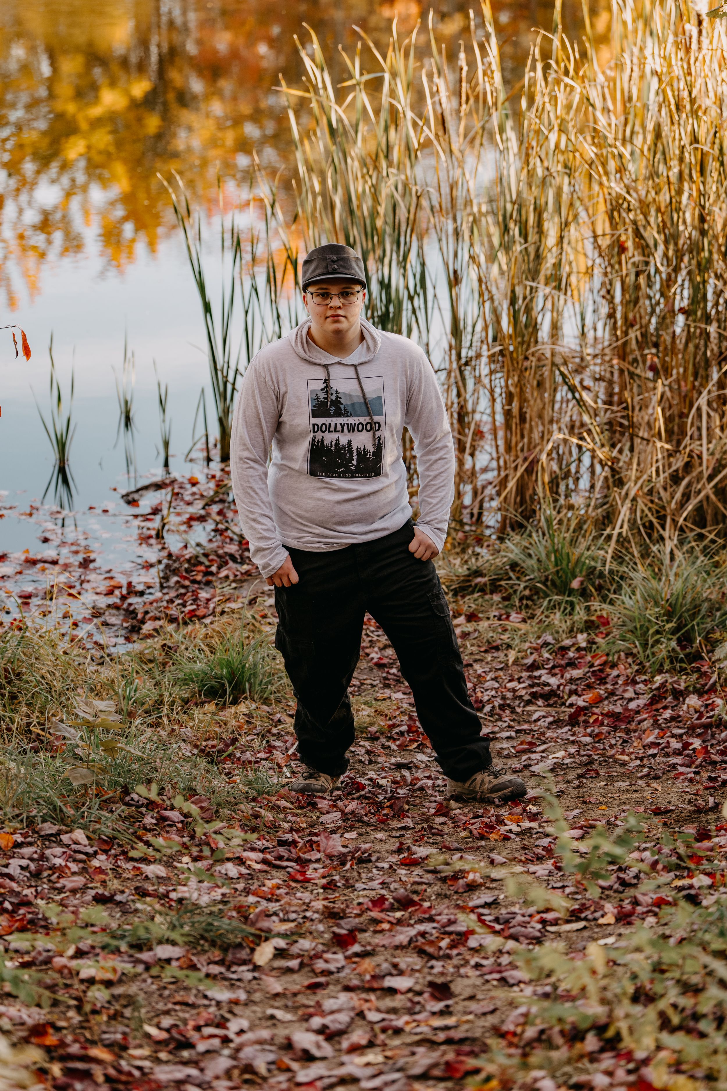 Person wearing light hoodie and cap standing by pond with autumn cattails and fallen leaves.