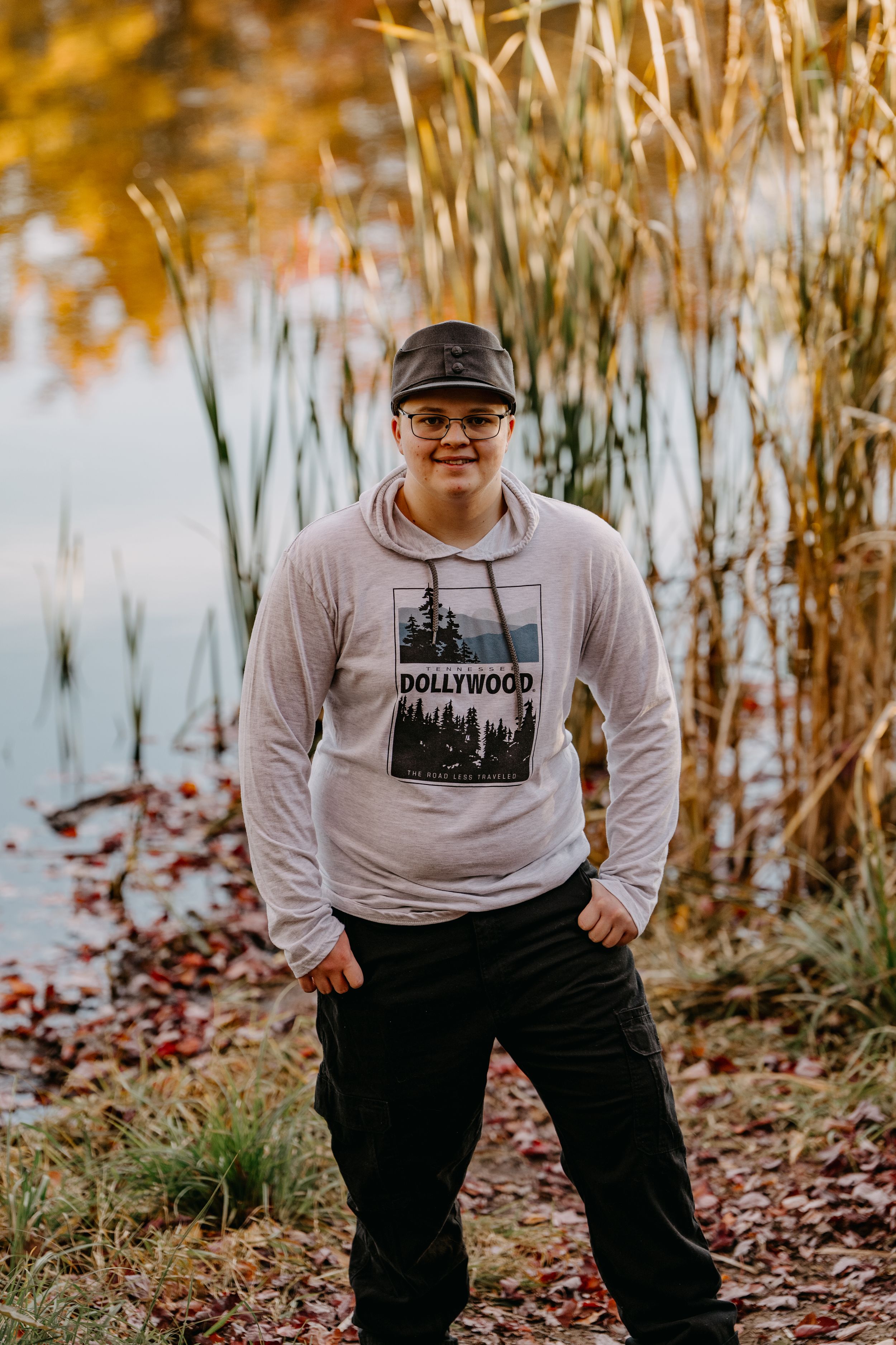 Person in a Hollywood graphic sweatshirt and baseball cap poses near autumn reeds by a pond with fall colors reflected in water.