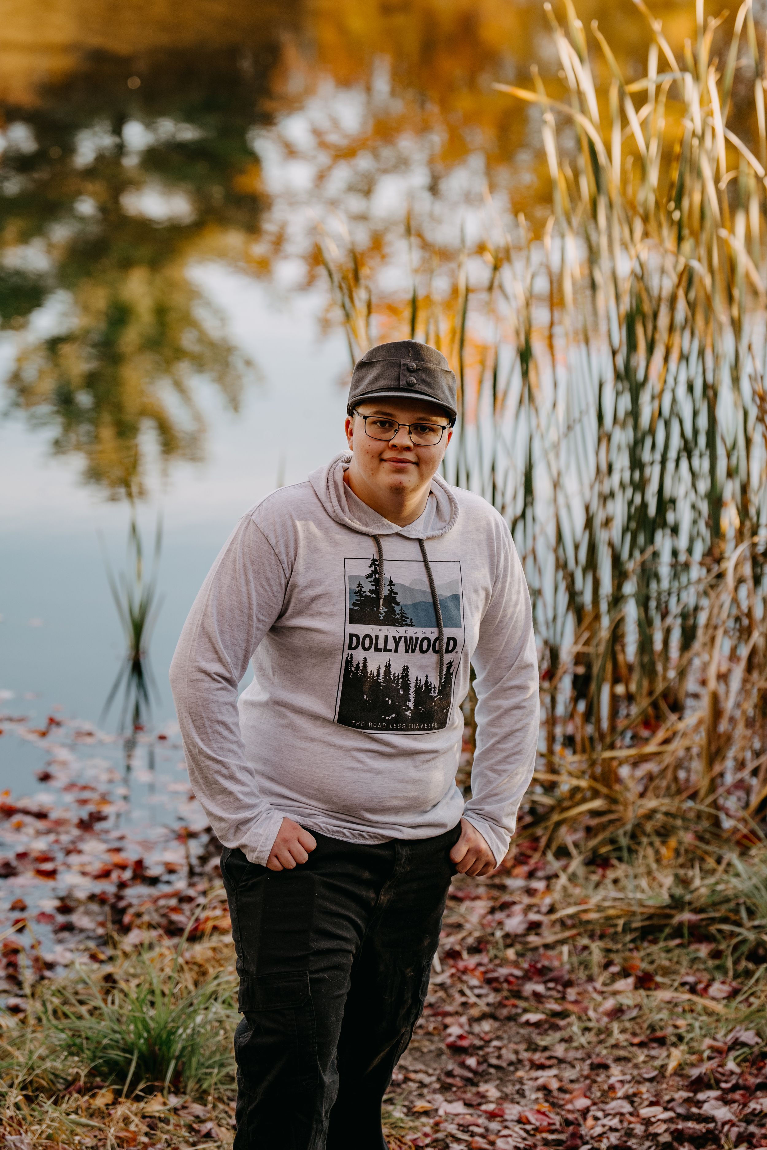 A person wearing a Hollywood hoodie poses by tall reeds near a lake during autumn.