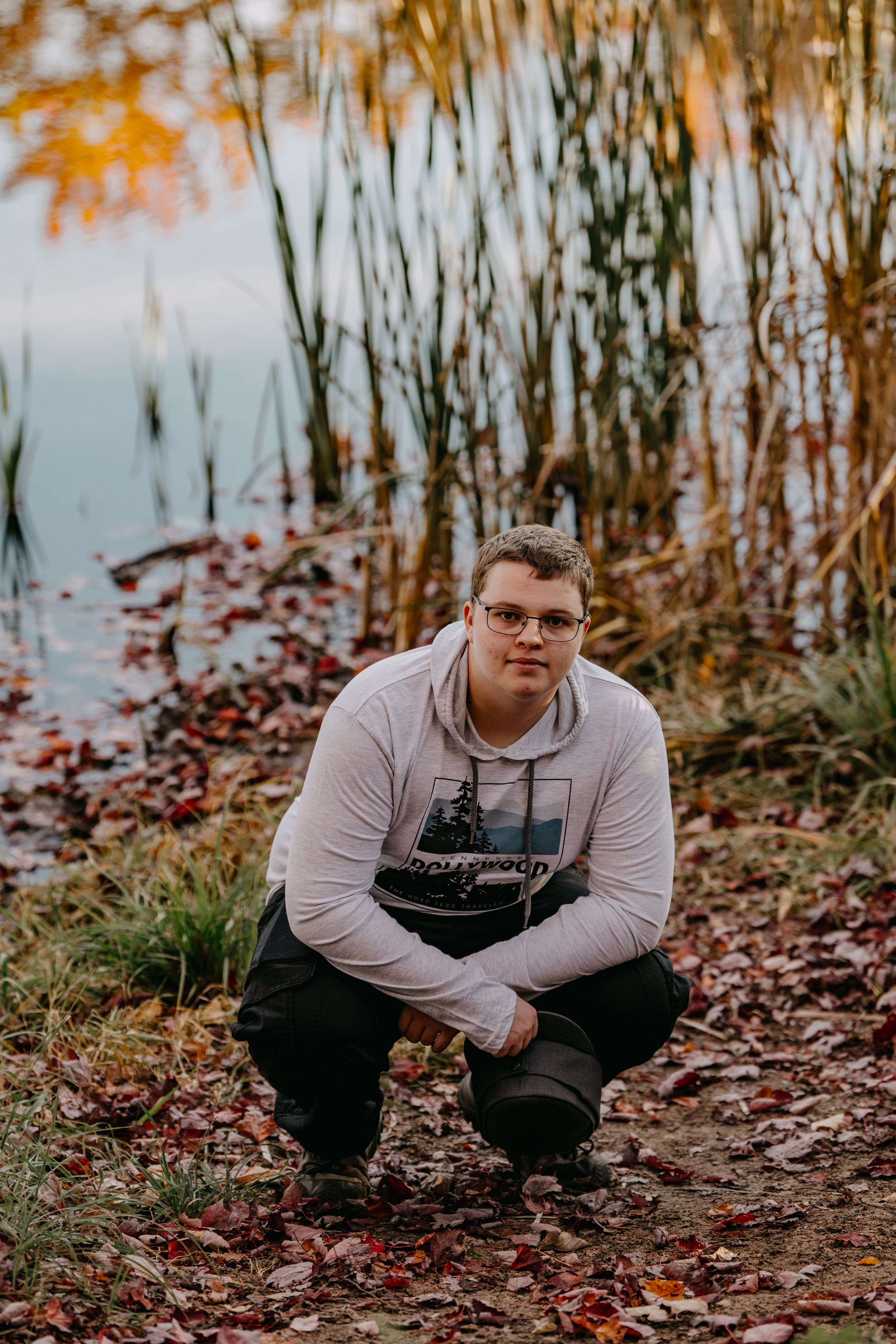 Someone poses in casual attire by a waterside with autumn leaves and reflective water.