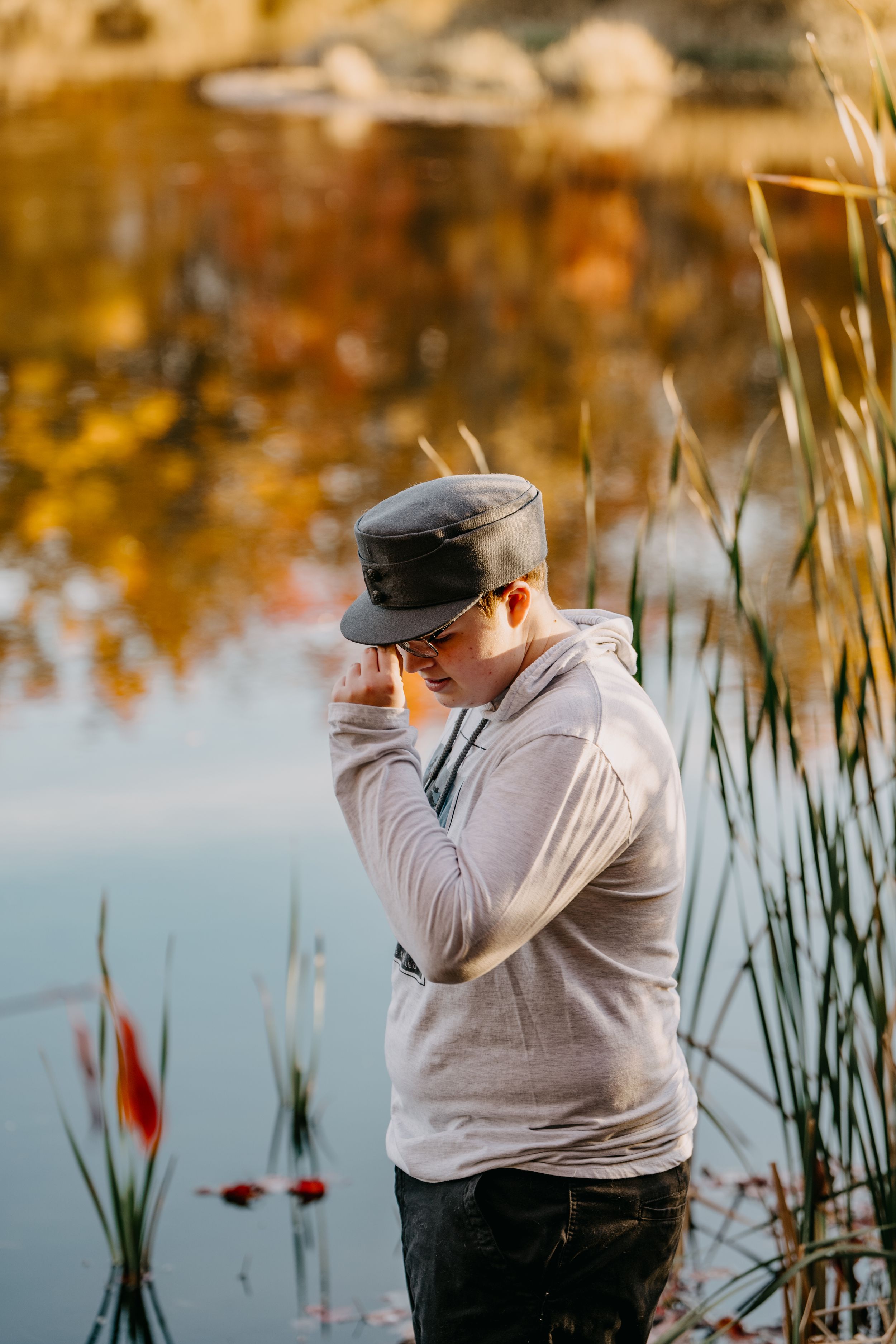 Person in light hoodie and cap photographing near pond with cattails at golden hour.