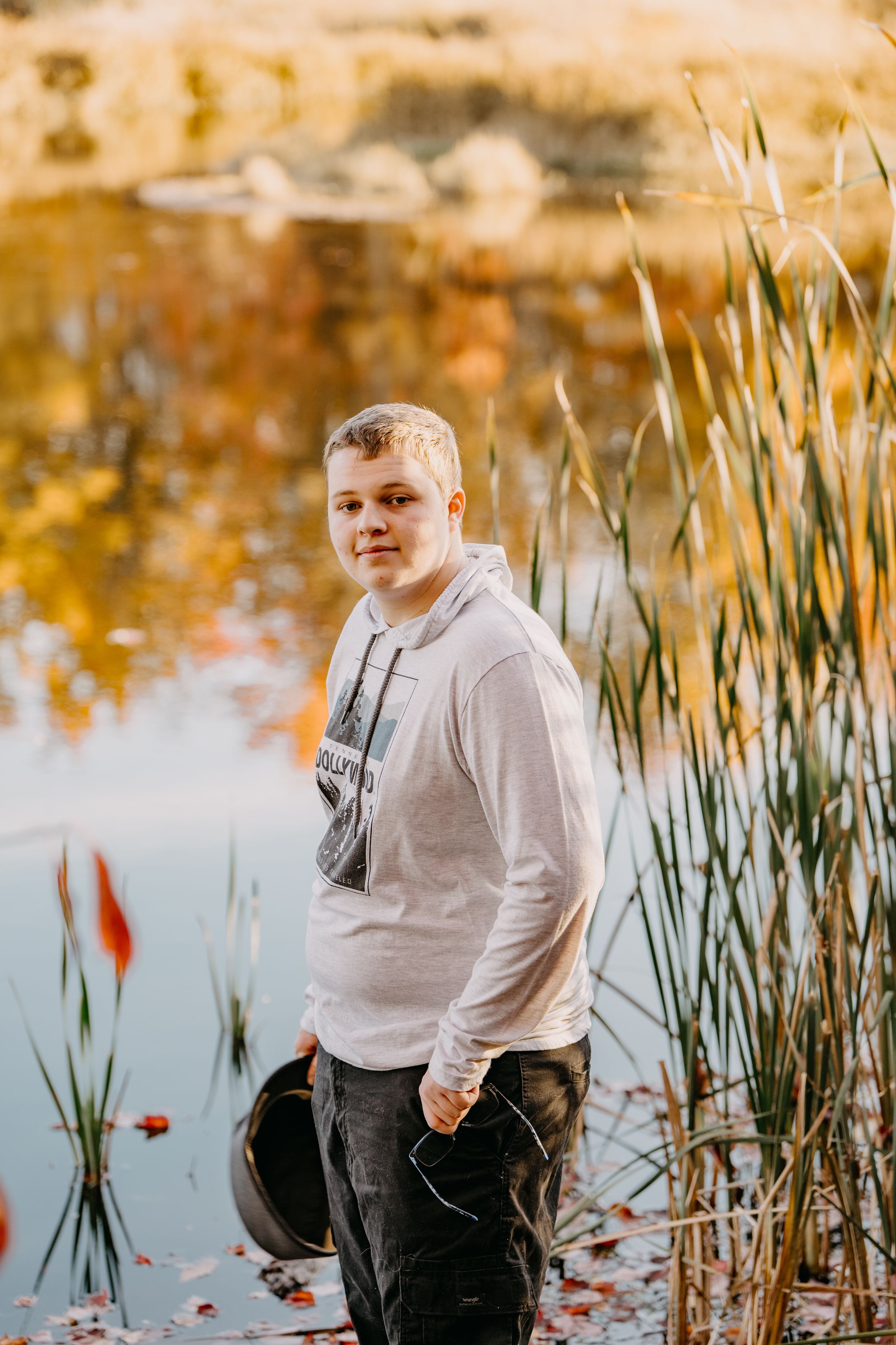 Someone poses in a light hoodie by a pond with cattails and autumn foliage reflected in the calm water.