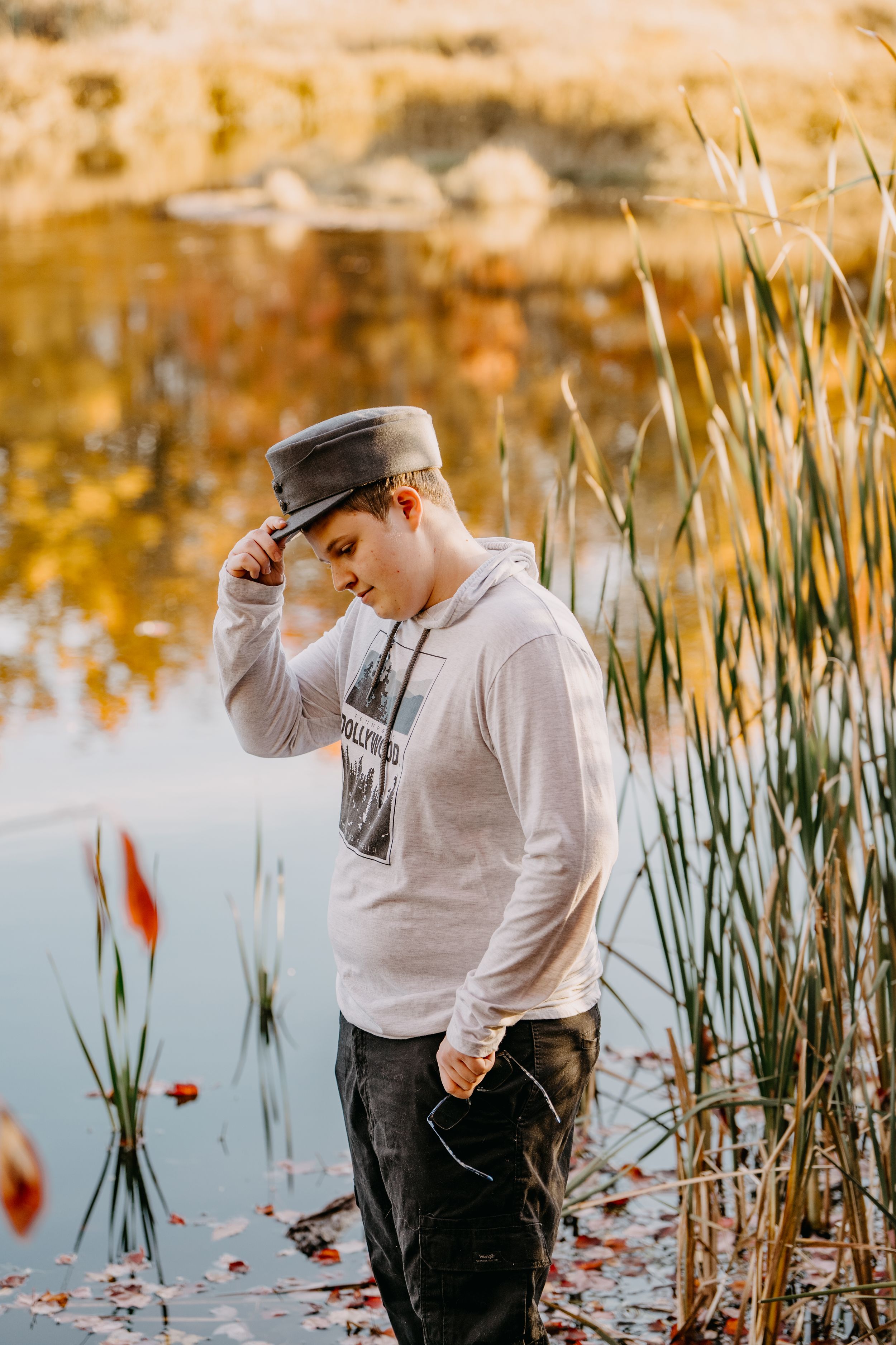A person in a gray hoodie and cap stands thoughtfully by a lake with golden reeds and fall reflections.