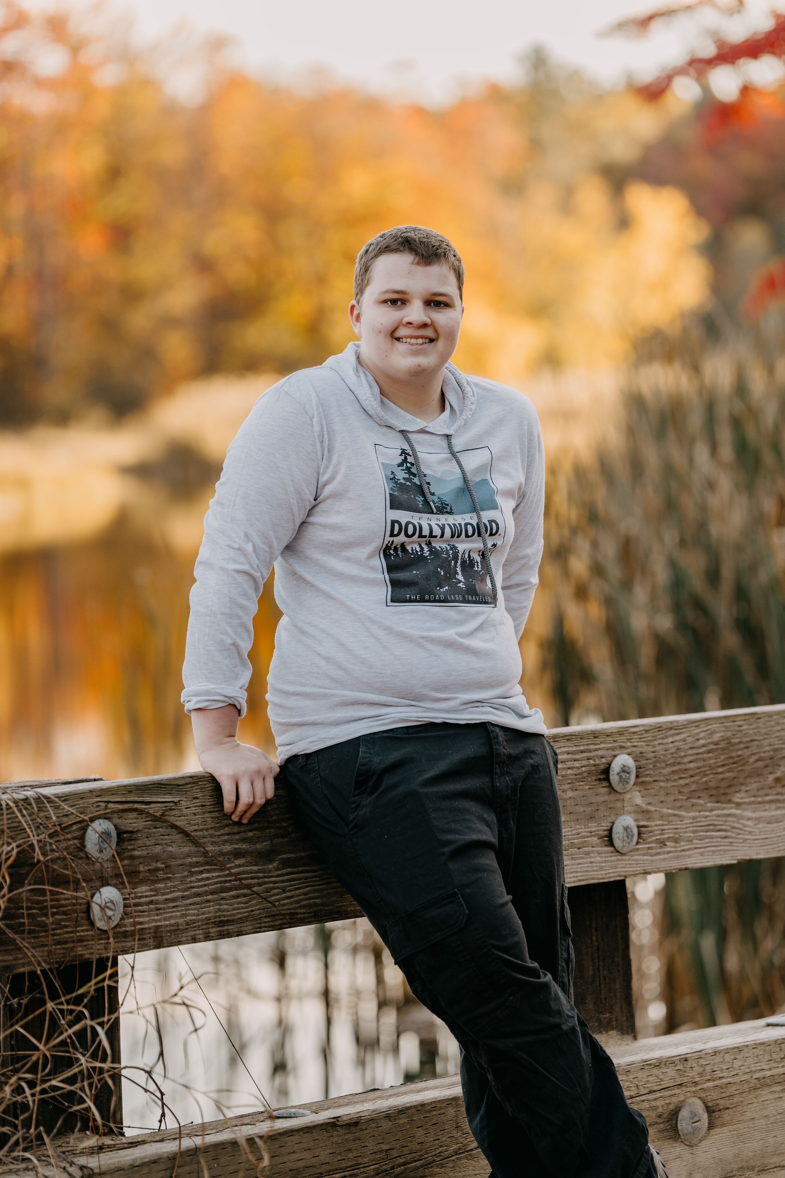 Person in graphic sweatshirt leaning on wooden railing with autumn reflections in water behind.