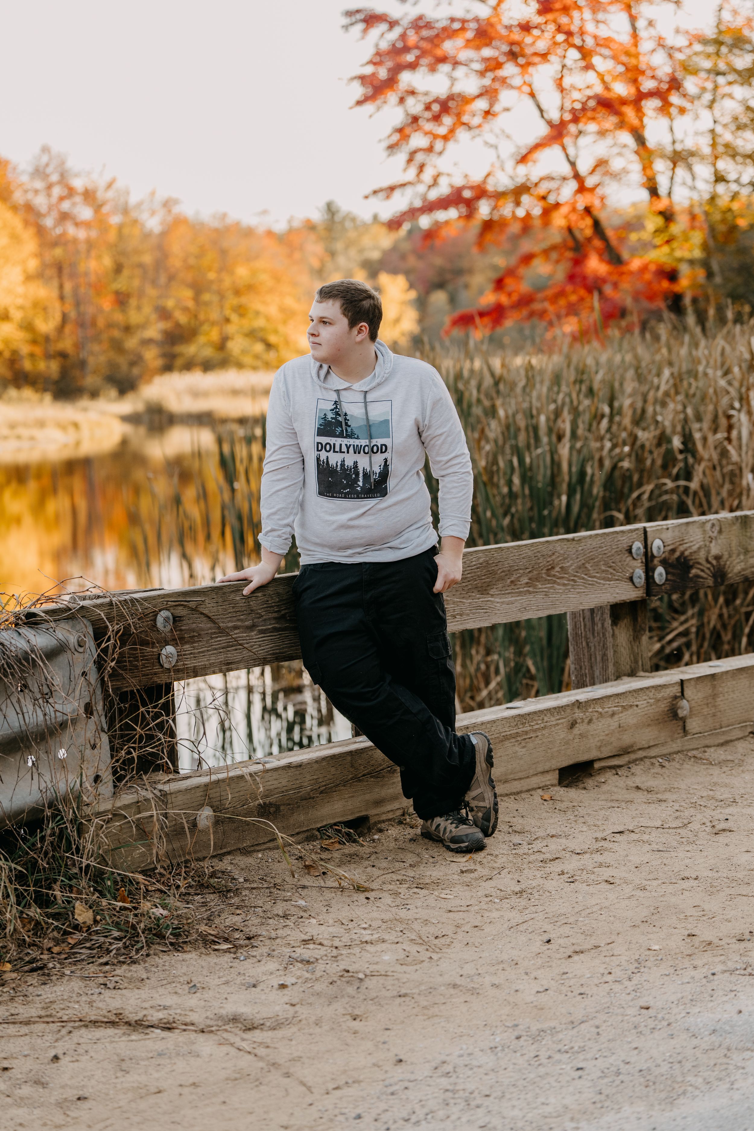 Person in a gray sweatshirt leans against wooden railing by autumn pond with colorful fall foliage.