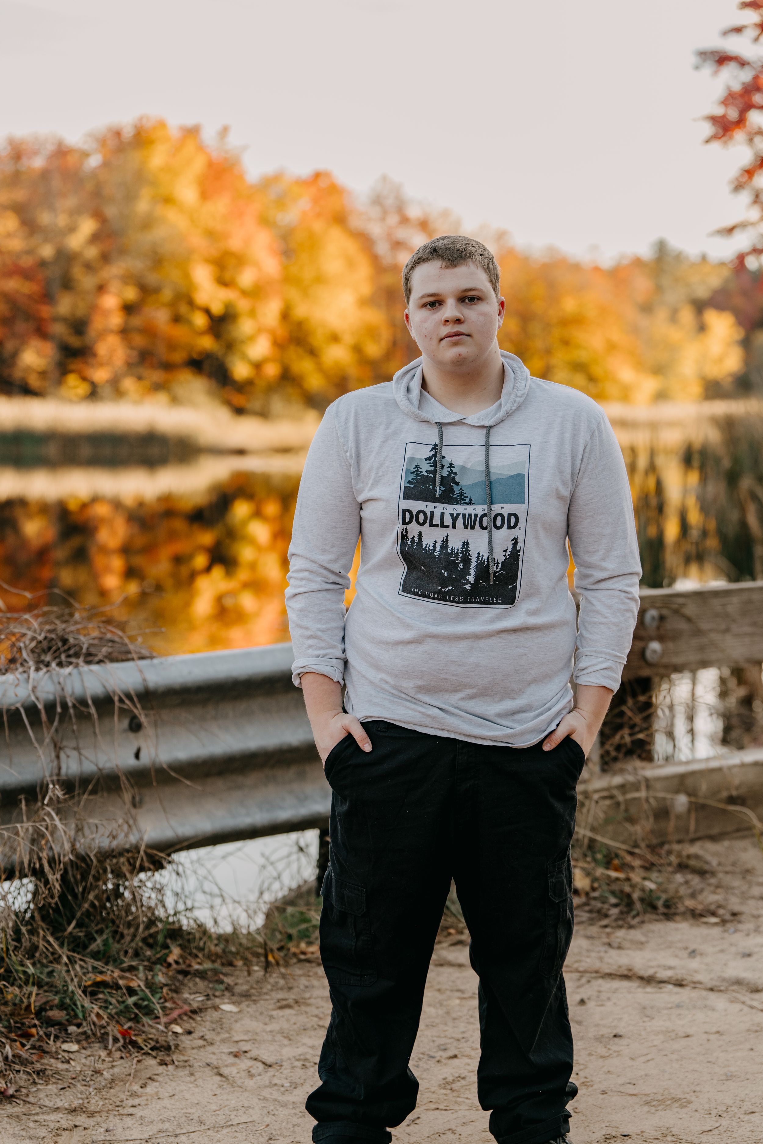 Person wearing Hollywood graphic hoodie stands on path by pond with autumn trees reflecting in water.